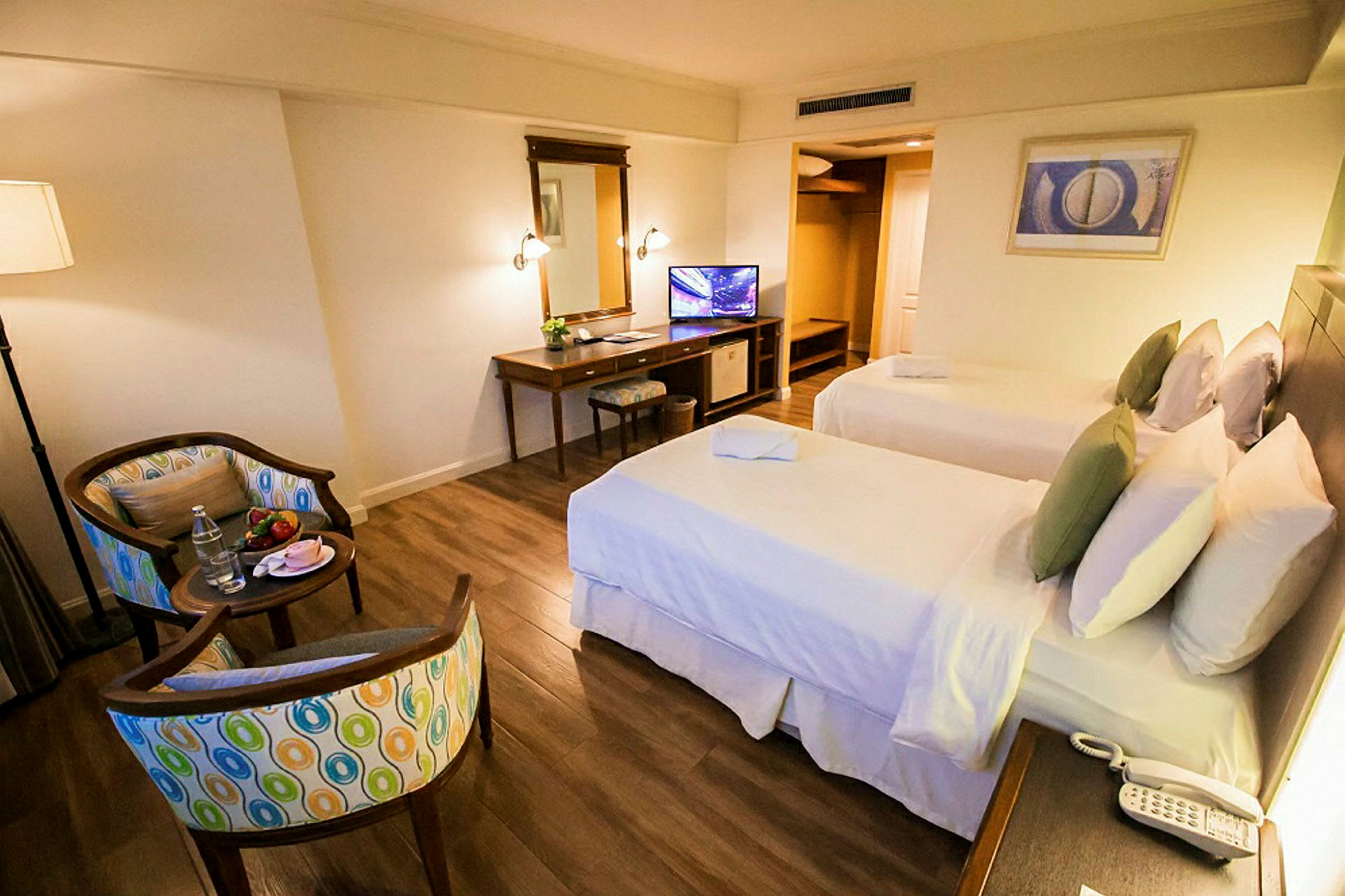 Ett hotellrum med två separata sängar, skrivbord, stol, spegel och en TV. Rummet har trägolv och en sittgrupp med två stolar och ett litet bord. (AI-genererad text)