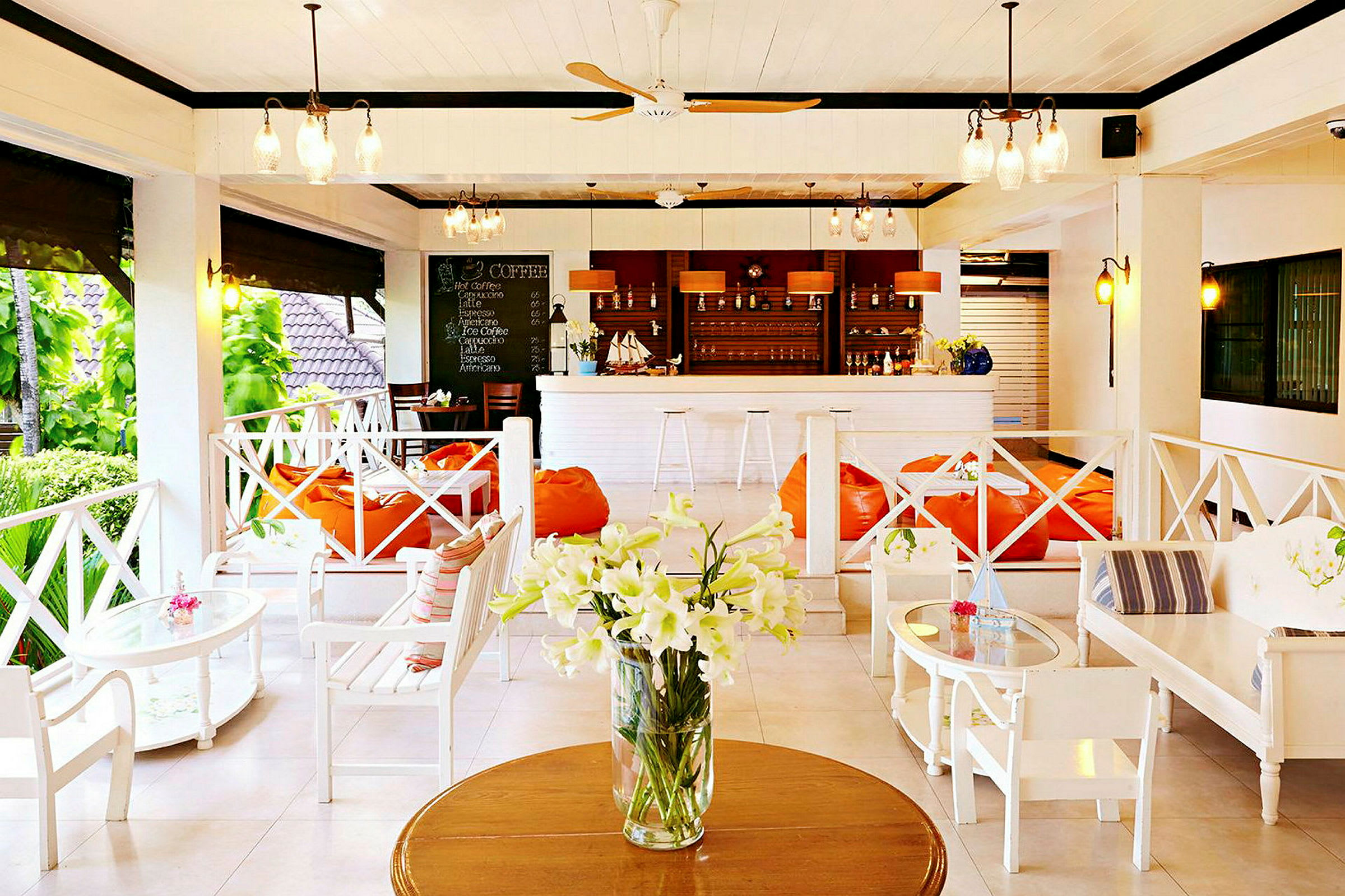 En ljus och öppen lounge med vita möbler, orange sittpuffar och en bar med drycker i bakgrunden samt en vas med vita blommor på ett bord. (AI-genererad text)