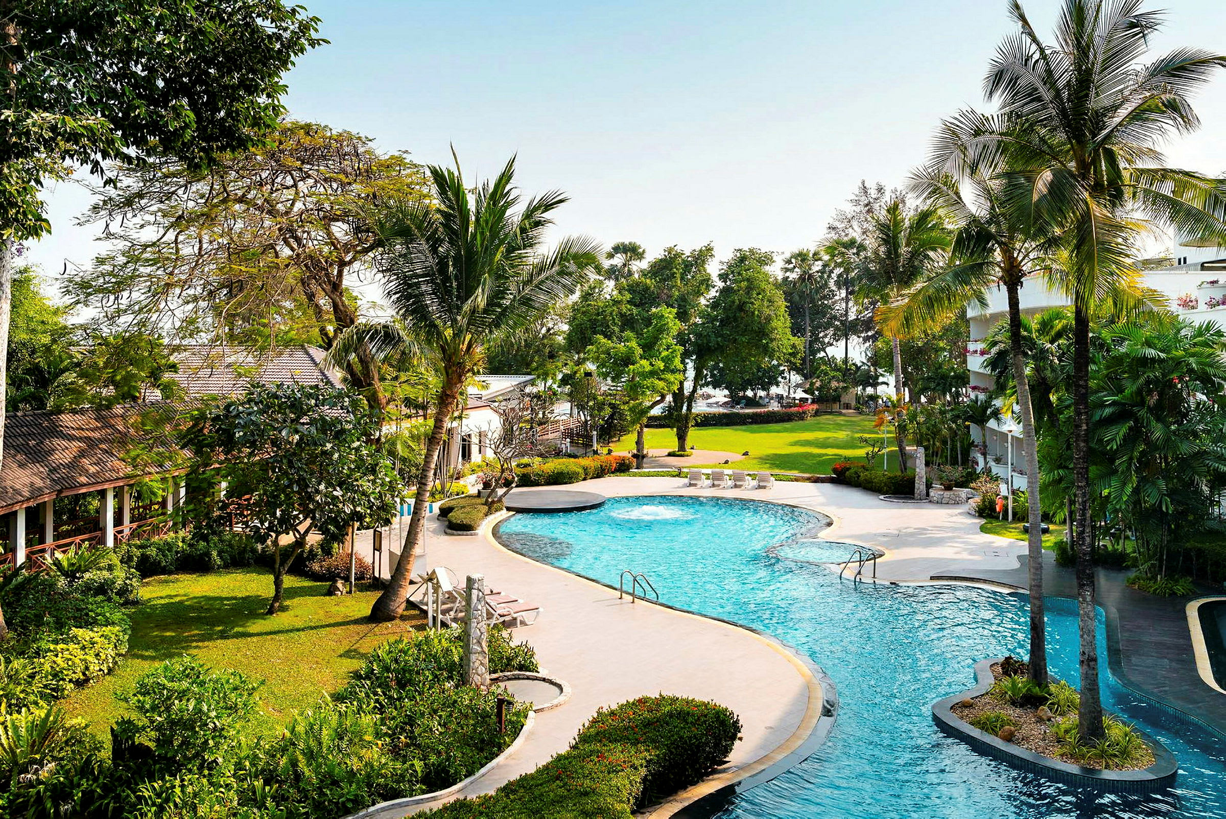 Ett hotellområde med en pool omgiven av grönska, palmer och byggnader. (AI-genererad text)
