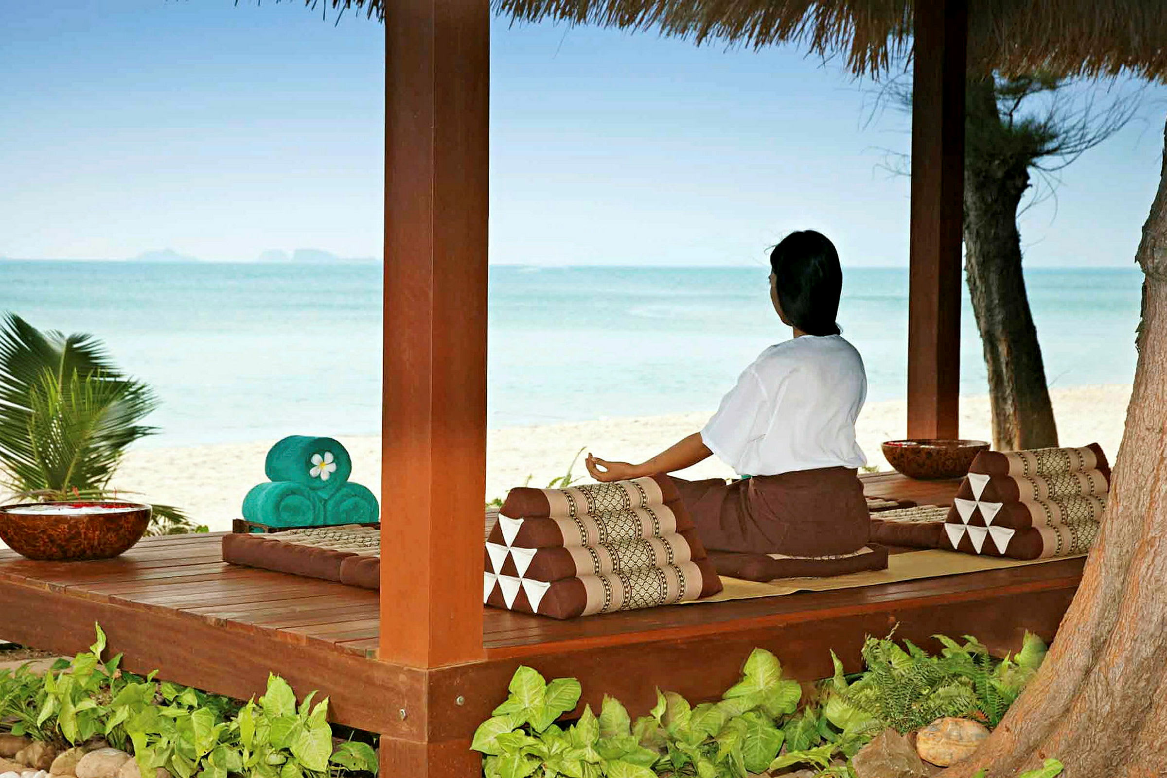 En person sitter i meditationsställning under ett träskjul med utsikt över stranden och havet. (AI-genererad text)