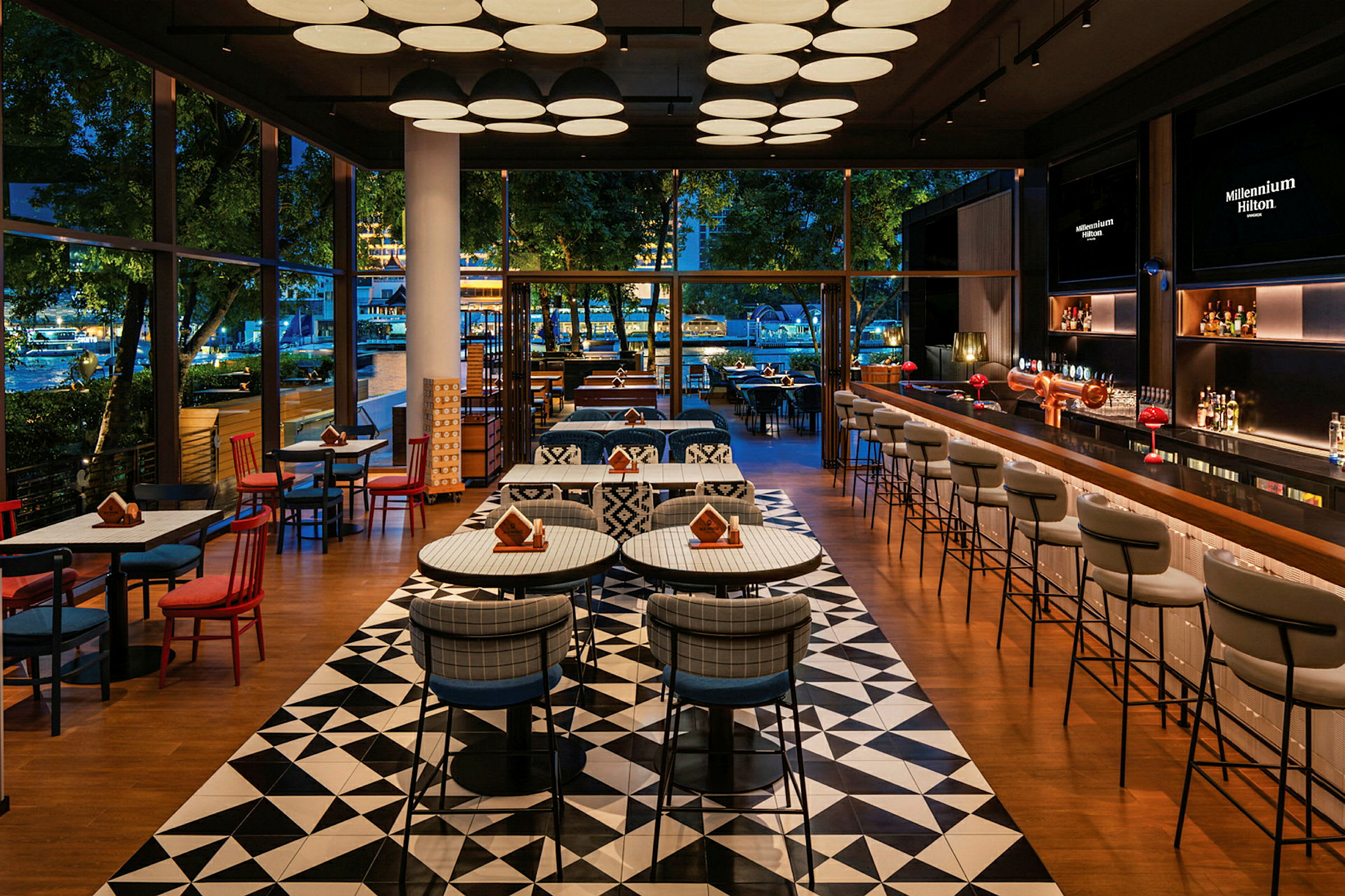 Ett elegant restaurangområde med moderna möbler, mönstrade golv, och stora glasfönster som visar utsikt mot en utomhusmiljö med träd. (AI-genererad text)