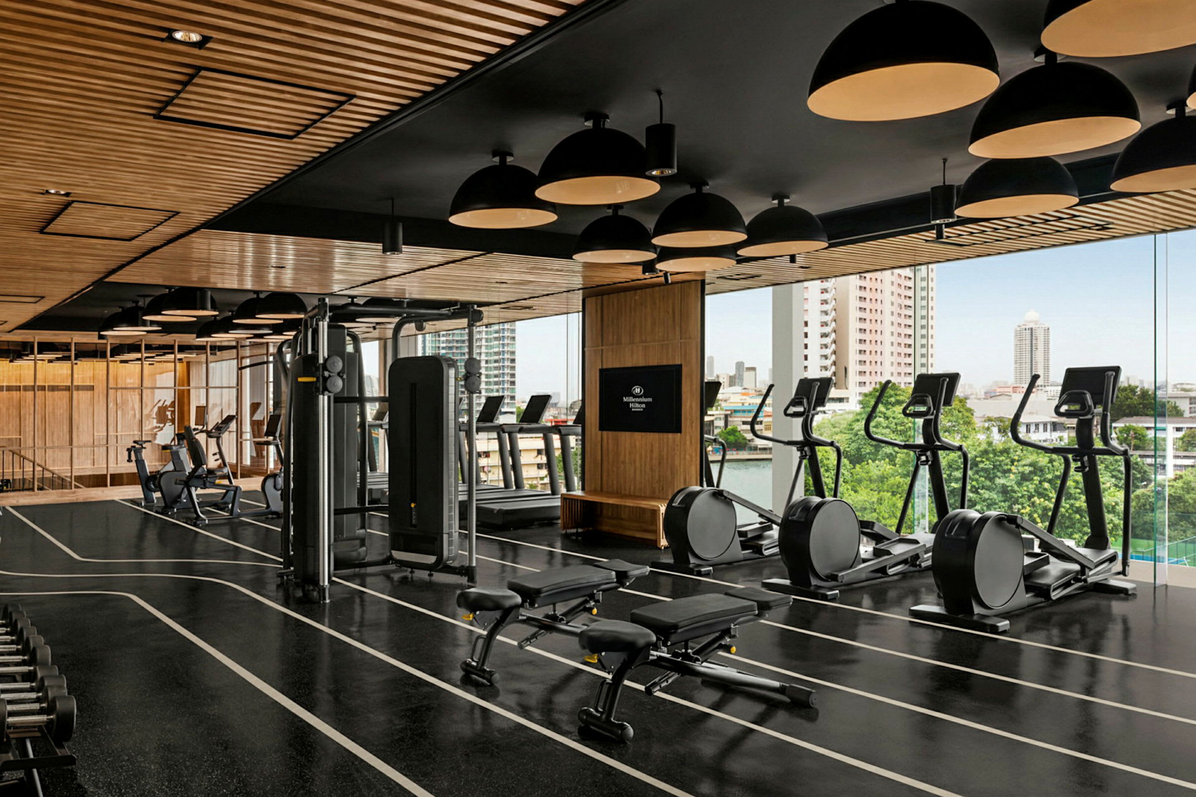 Ett välutrustat gym med träningsmaskiner, fria vikter och bänkar. Lokalen är ljus med stora fönster som erbjuder utsikt över staden. (AI-genererad text)