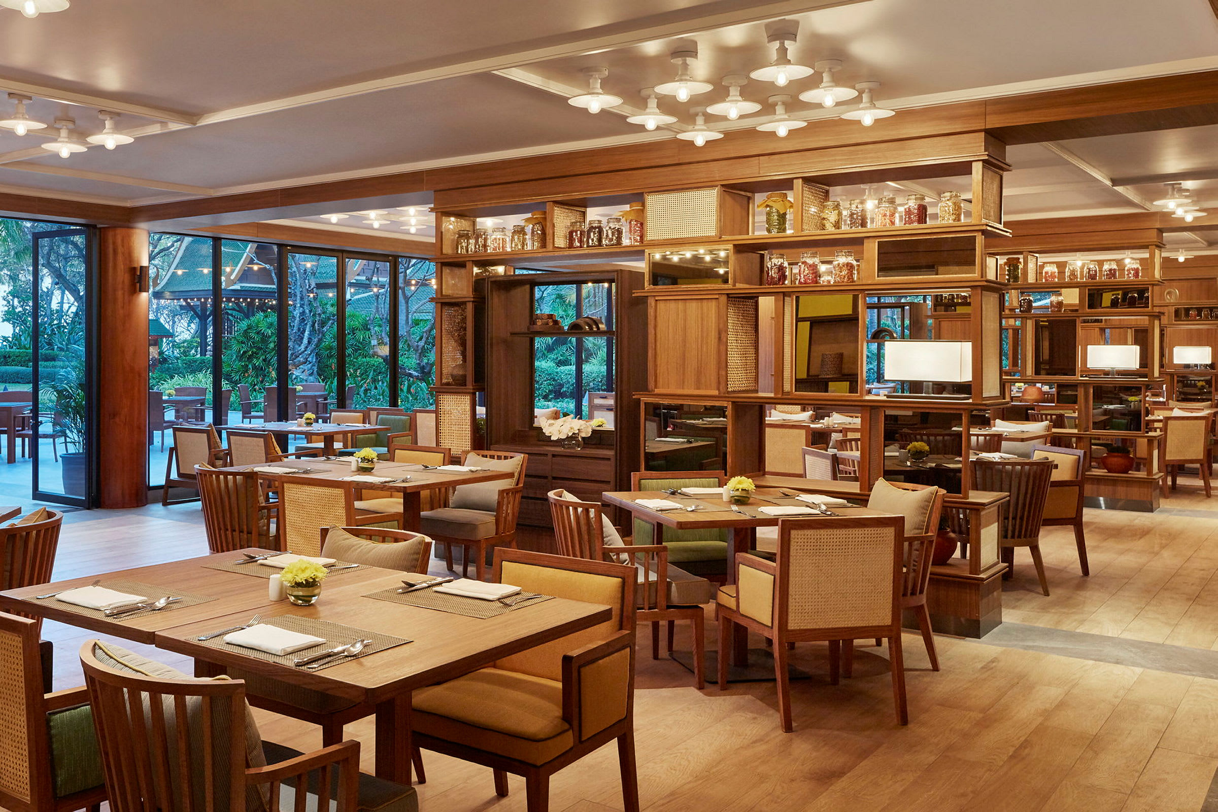 En elegant restaurang med träinredning, bord och stolar arrangerade i en öppen sal. Stora fönster visar utsikten mot grönska utomhus. (AI-genererad text)