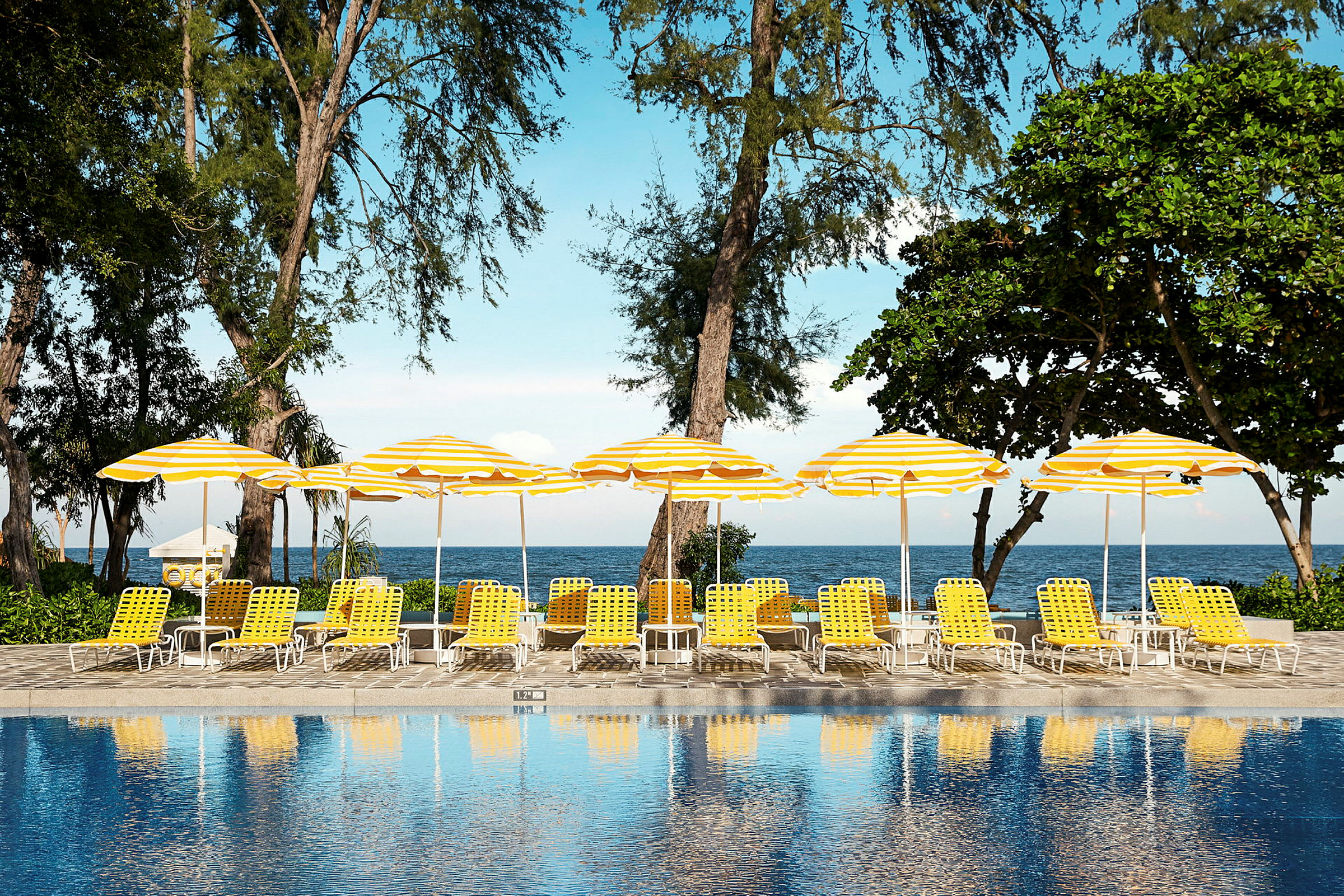 En pool framför solstolar med gula och vita parasoller nära stranden. (AI-genererad text)
