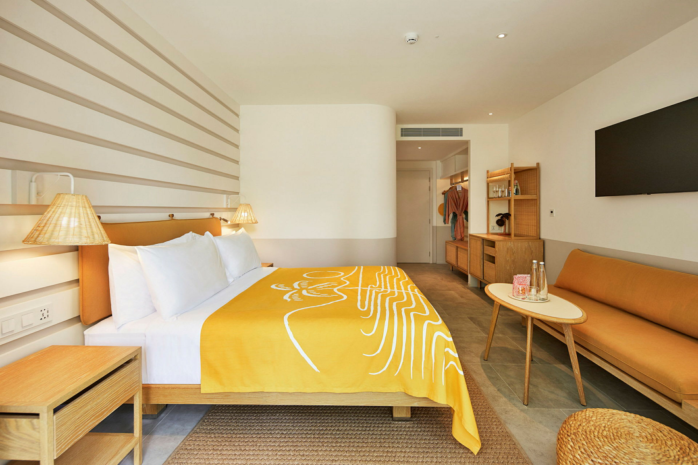 Ett modernt hotellrum med en säng täckt av en gul filt, en soffa, ett bord och en stor TV på väggen. (AI-genererad text)