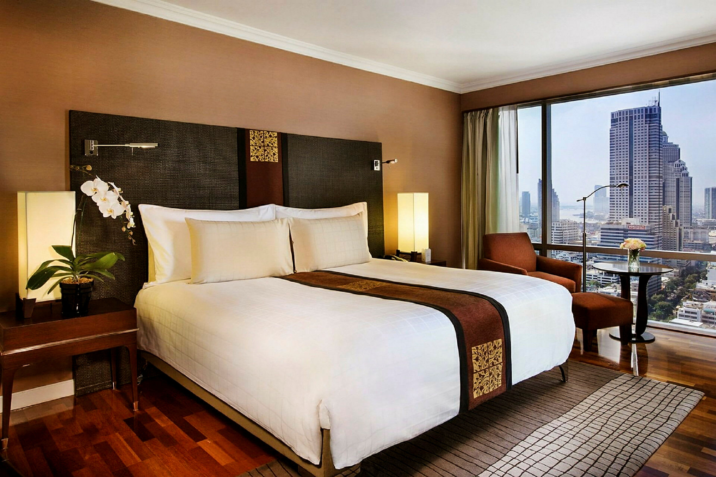 Ett hotellrum med en stor dubbelsäng, dekorerad med en elegant brun rand och guldmönster. Rummet har moderna möbler och stora fönster med utsikt över en stad. (AI-genererad text)