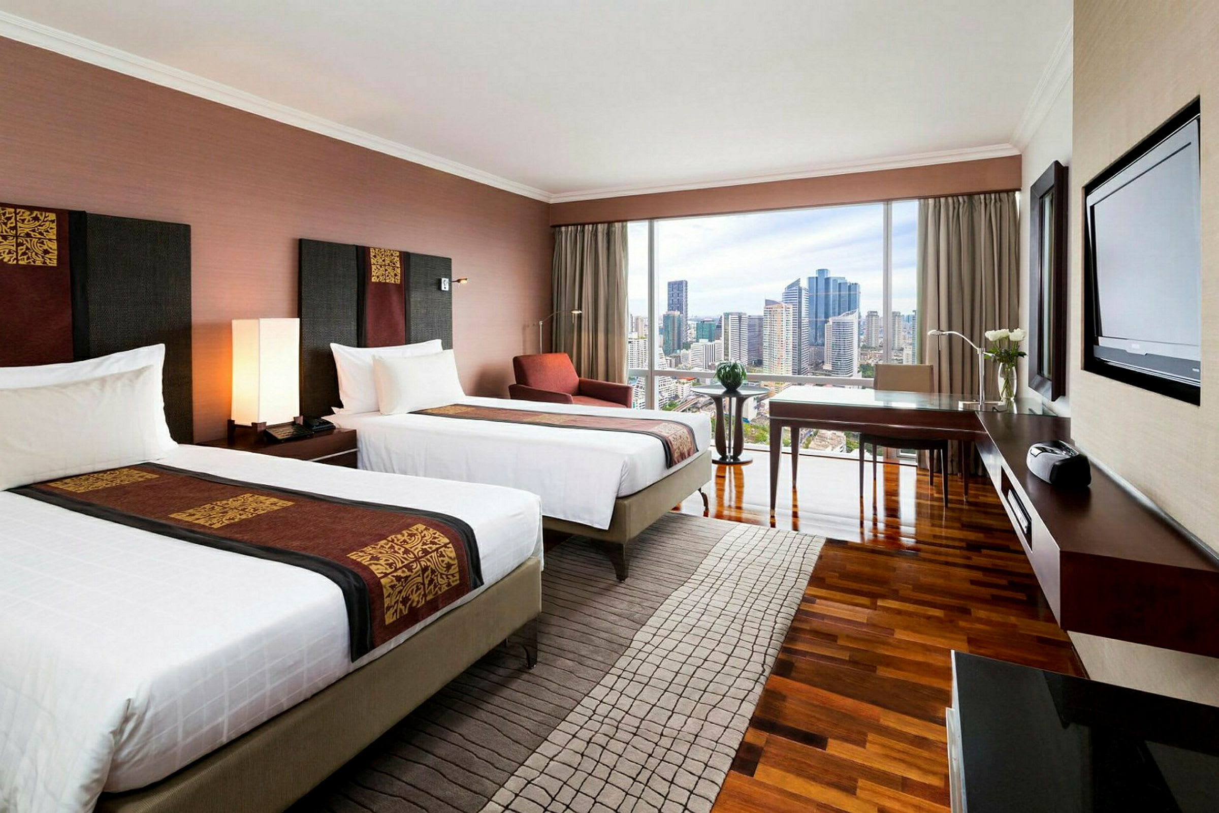 Ett modernt hotellrum med två enkelsängar, ett skrivbord, en stol och stora fönster med utsikt över stadens skyline. (AI-genererad text)