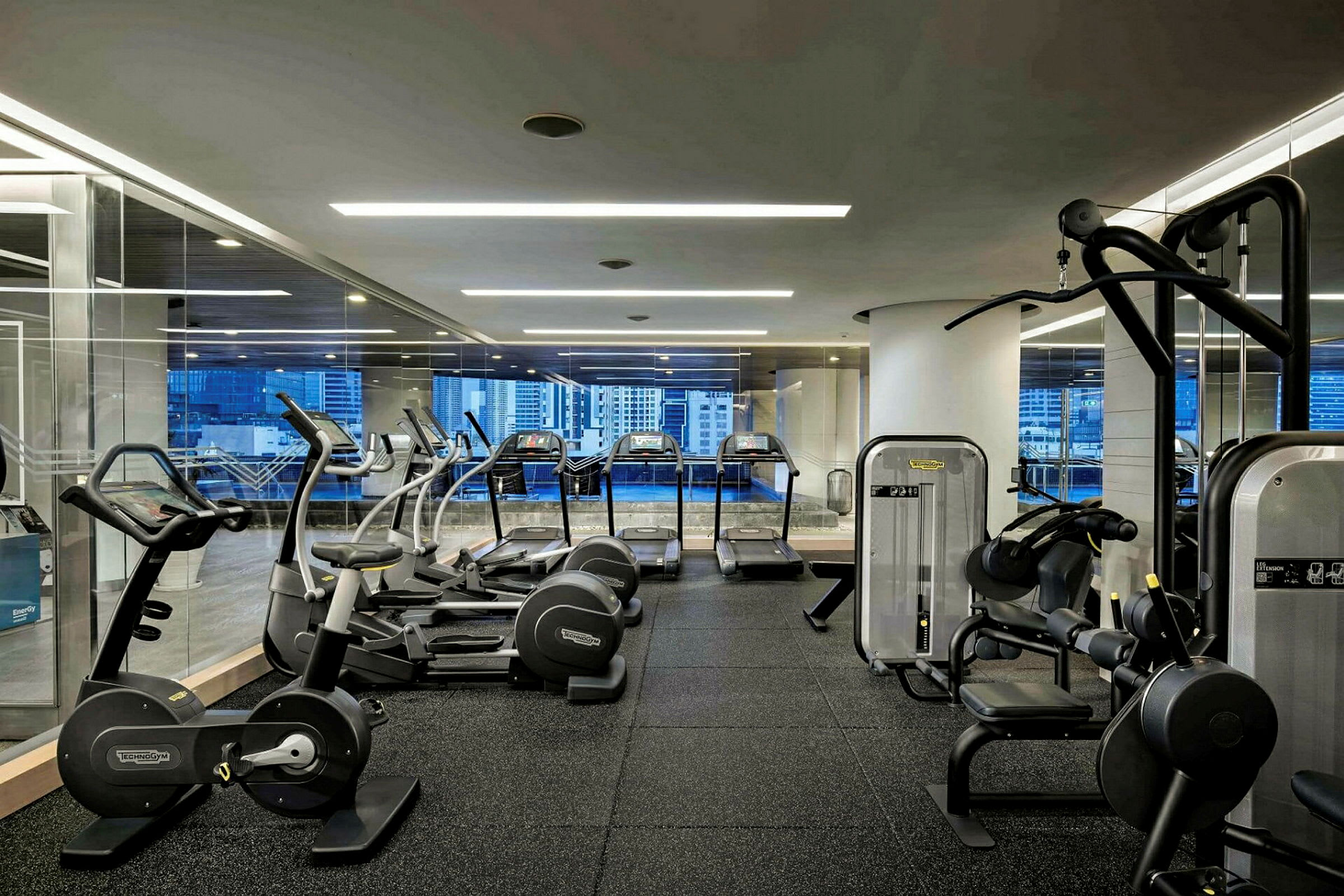 Ett gym med olika träningsmaskiner såsom löpband, motionscyklar och styrketräningsmaskiner. Stora fönster visar en utsikt av stadsmiljö. (AI-genererad text)