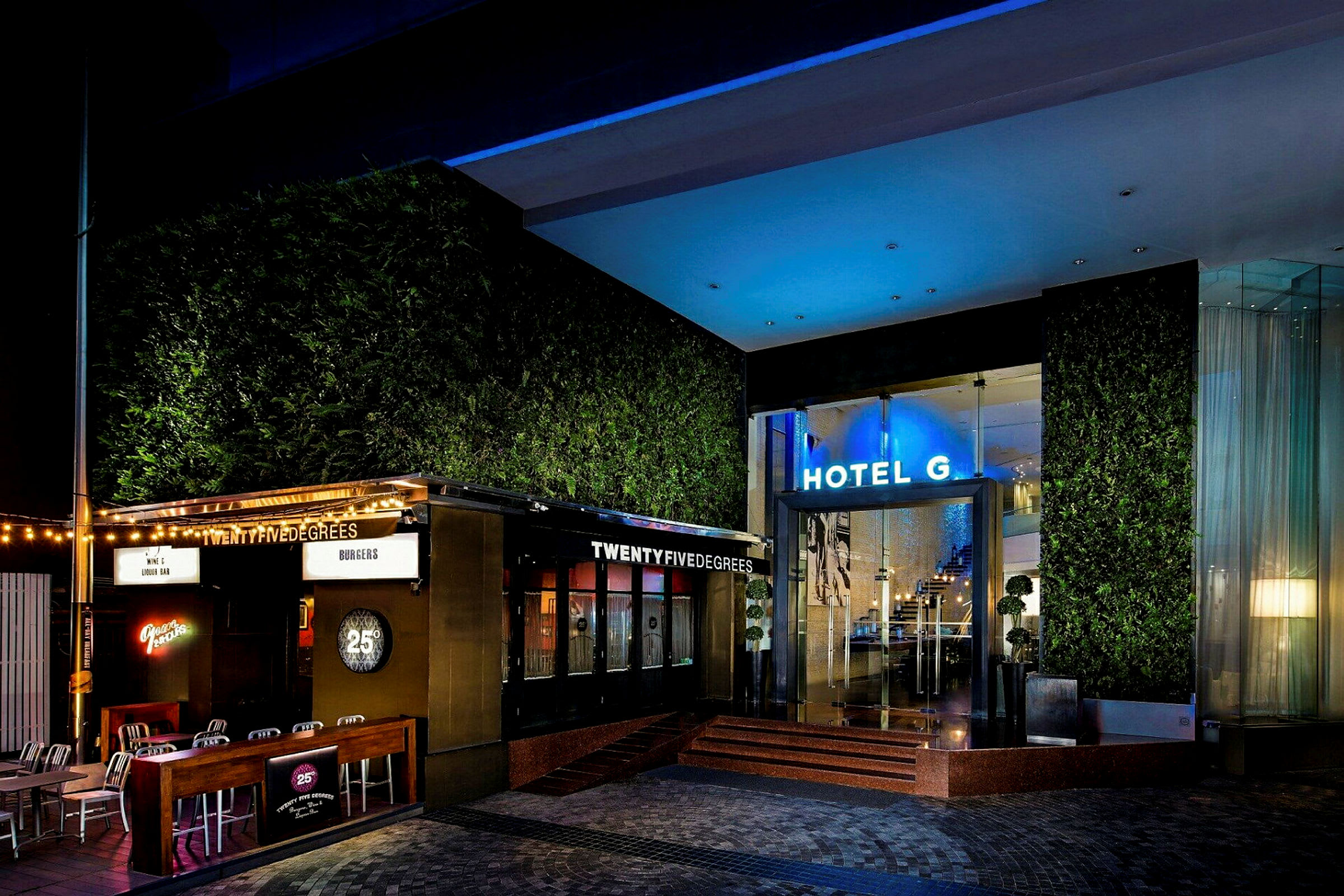 En upplyst hotellentré med en restaurang bredvid, omgiven av grönska och moderna detaljer. (AI-genererad text)