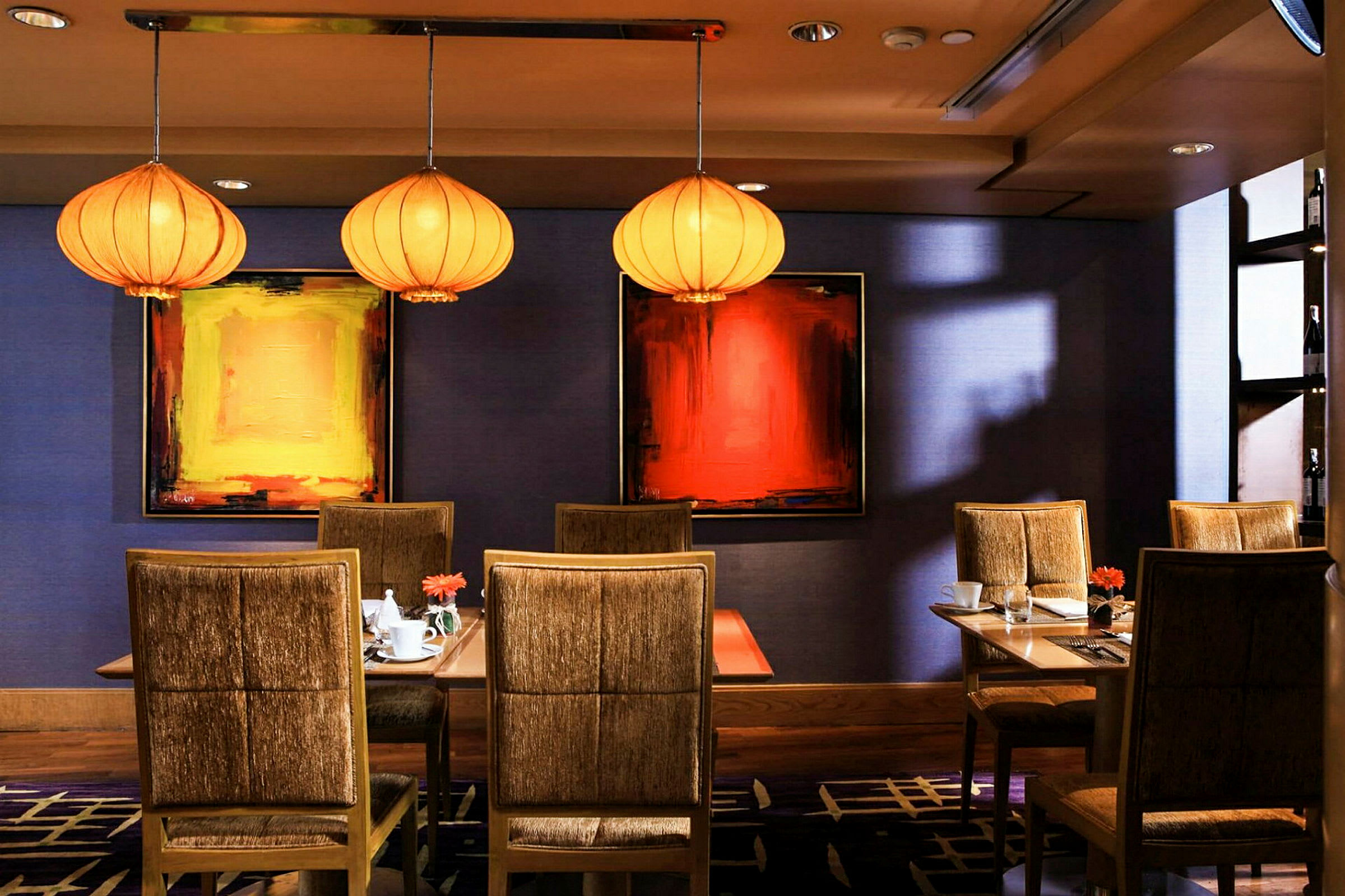 En interiörbild av en restaurang med tre bord omgivna av stoppade stolar, samt konst på väggarna och dekorativa lampor i taket. (AI-genererad text)