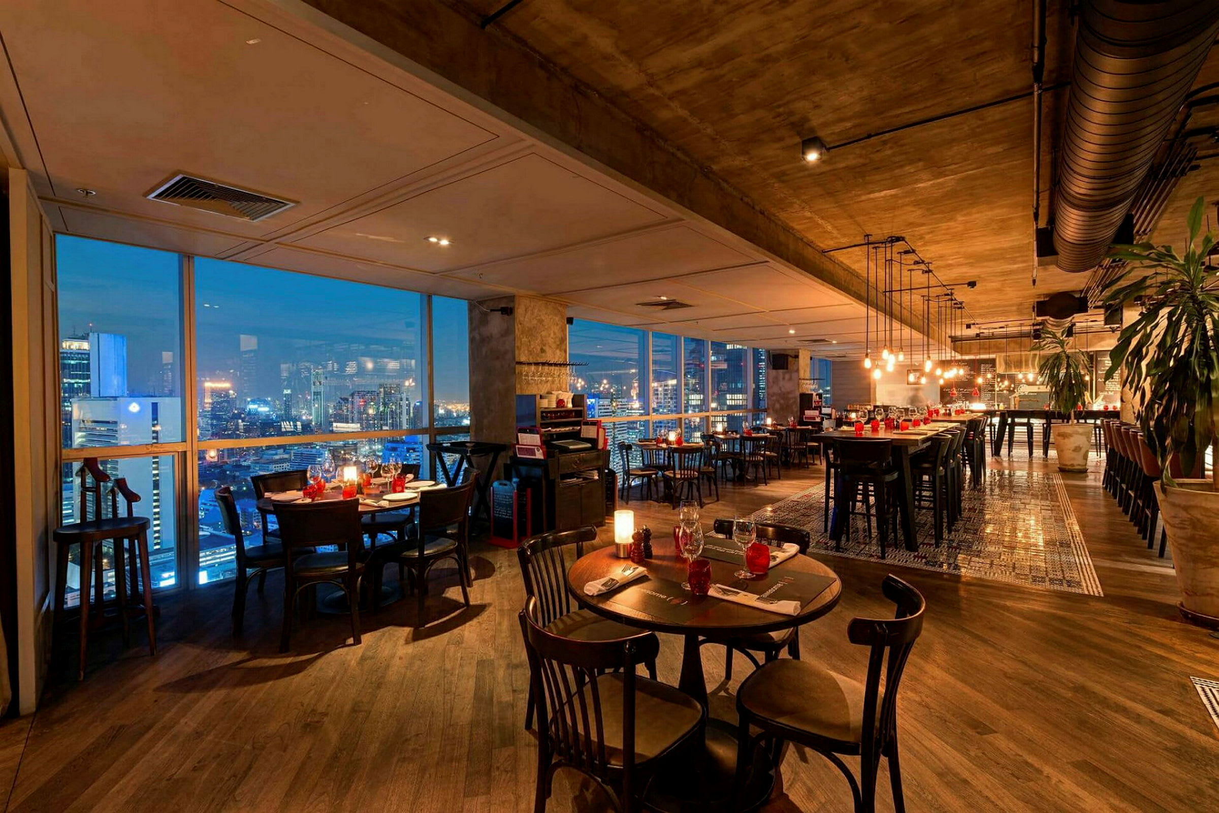 En elegant restaurang med moderna möbler, fönster som visar en stadsutsikt på kvällen och belysning som skapar en mysig atmosfär. (AI-genererad text)