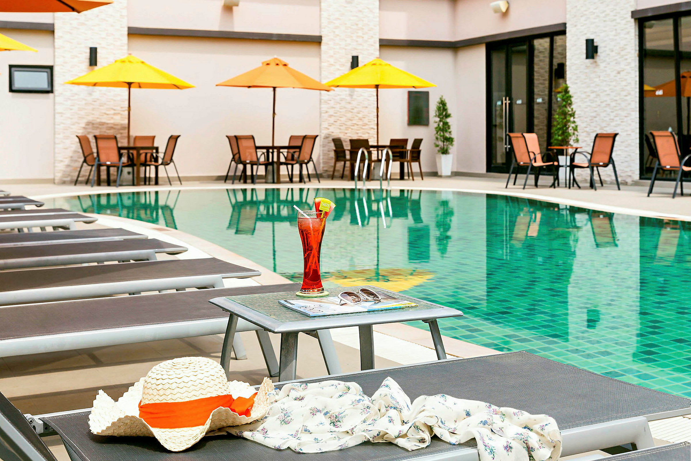 En pool med solstolar och ett bord i förgrunden som har en sommarhatt, en dryck och solglasögon på sig. (AI-genererad text)