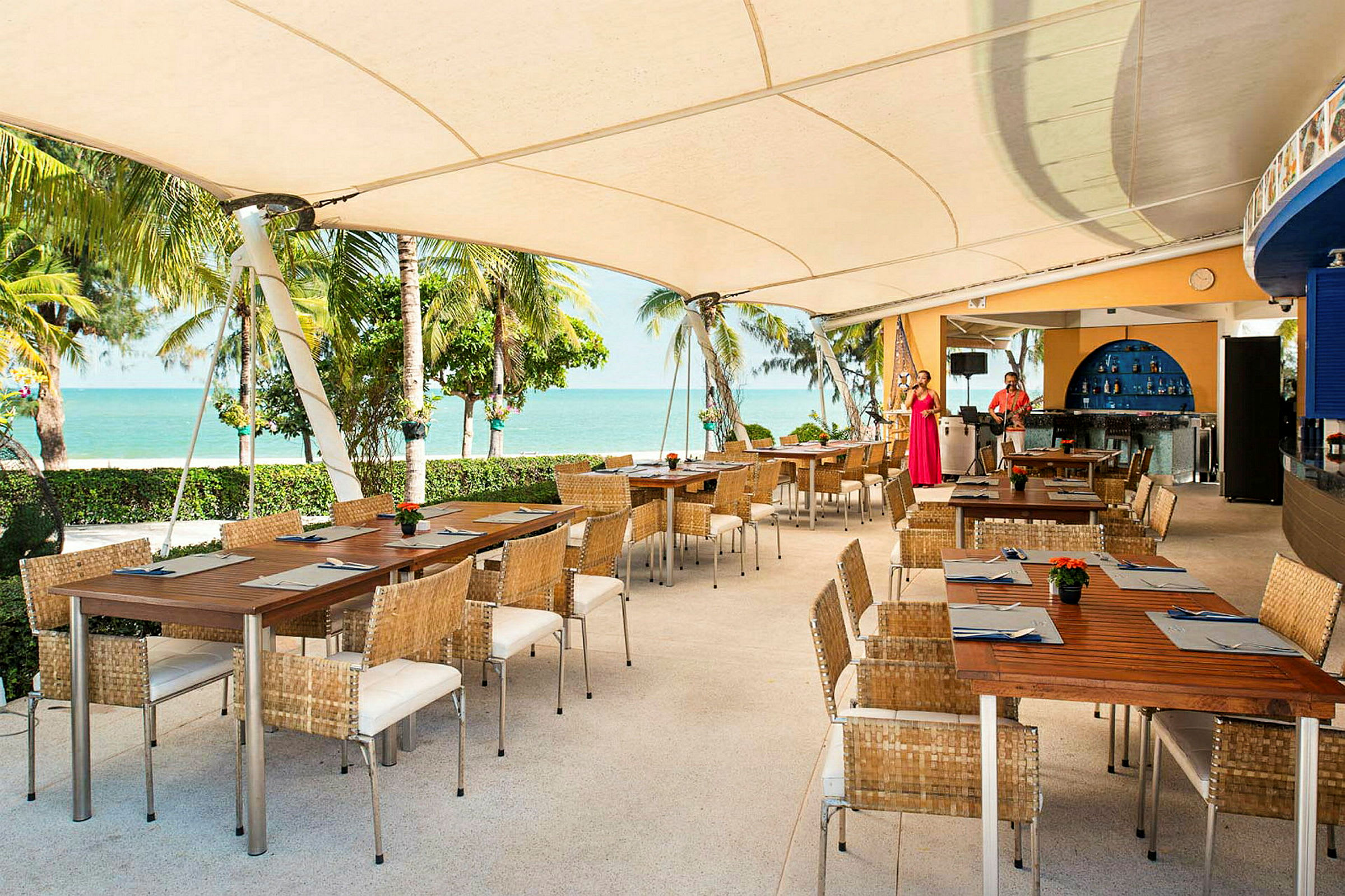 En utomhusrestaurang med träbord och korgstolar under ett stort tak. Utsikt över stranden och havet med palmer i bakgrunden. (AI-genererad text)
