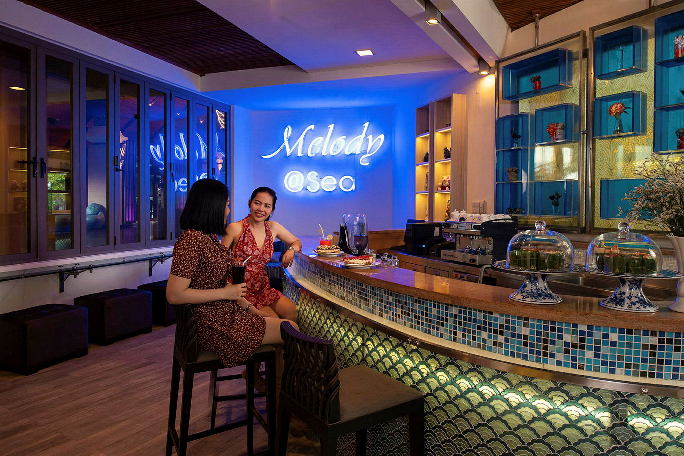 Två personer sitter vid en bardisk på en lokal med namnet 'Melody @Sea' skrivet i neon på väggen. Inredningen är stilfull med färgstarka dekorationer. (AI-genererad text)