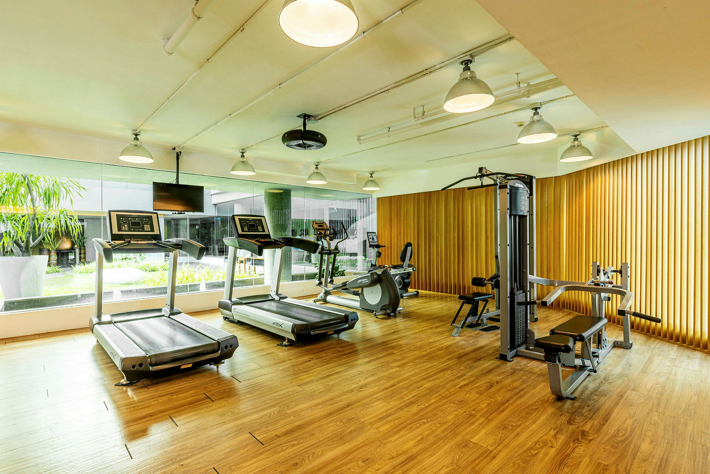 Ett gym med löpband, motionscykel och styrketräningsutrustning i ett rum med trägolv och fönster mot en grön uteplats. (AI-genererad text)