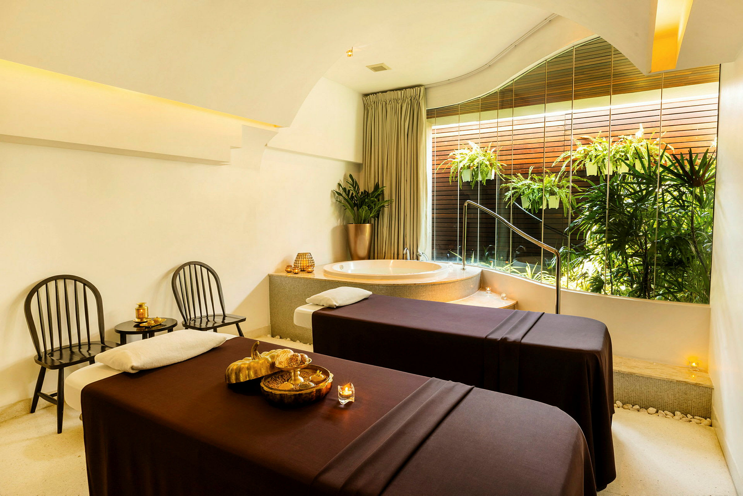 Ett spa-rum med två massagebänkar, en jacuzzi och växter vid ett stort fönster. (AI-genererad text)