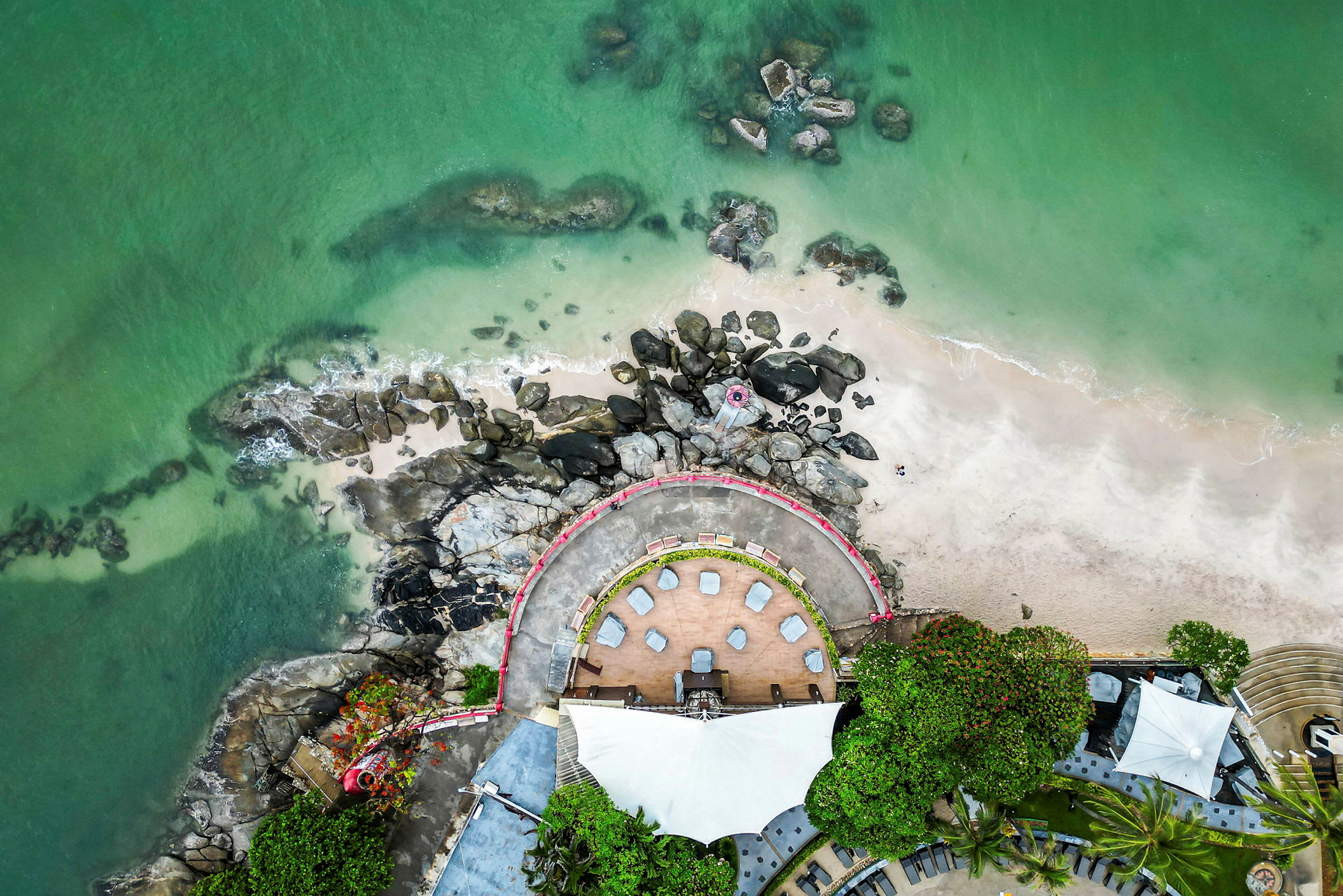 Flygfoto av en strand med grönt vatten, stenar samt en uteservering med sittplatser och träd. (AI-genererad text)