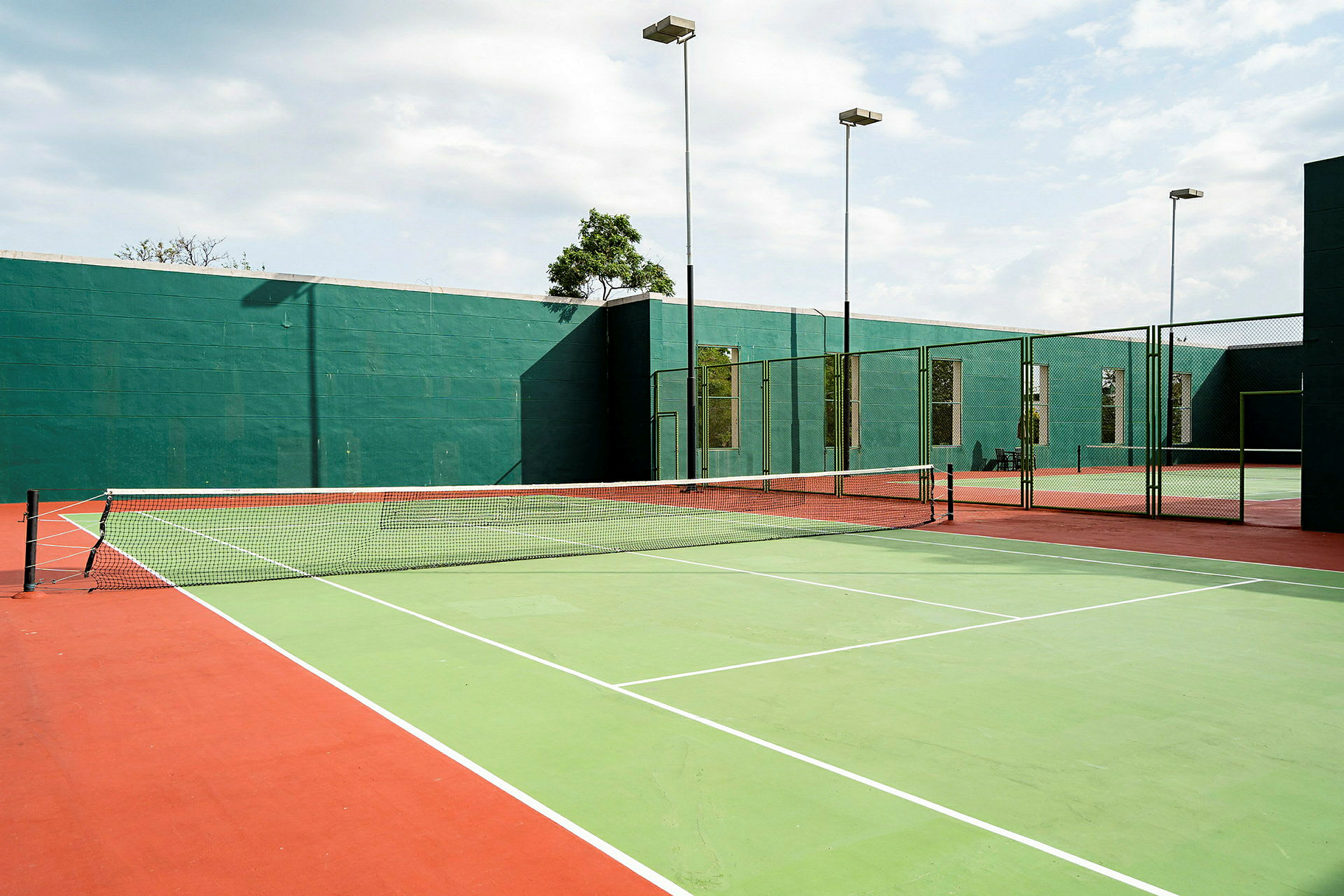 Tennisbanor utomhus med gröna och röda markeringar samt nät i mitten. Bakom banorna syns gröna väggar och höga stolpar med lampor. (AI-genererad text)