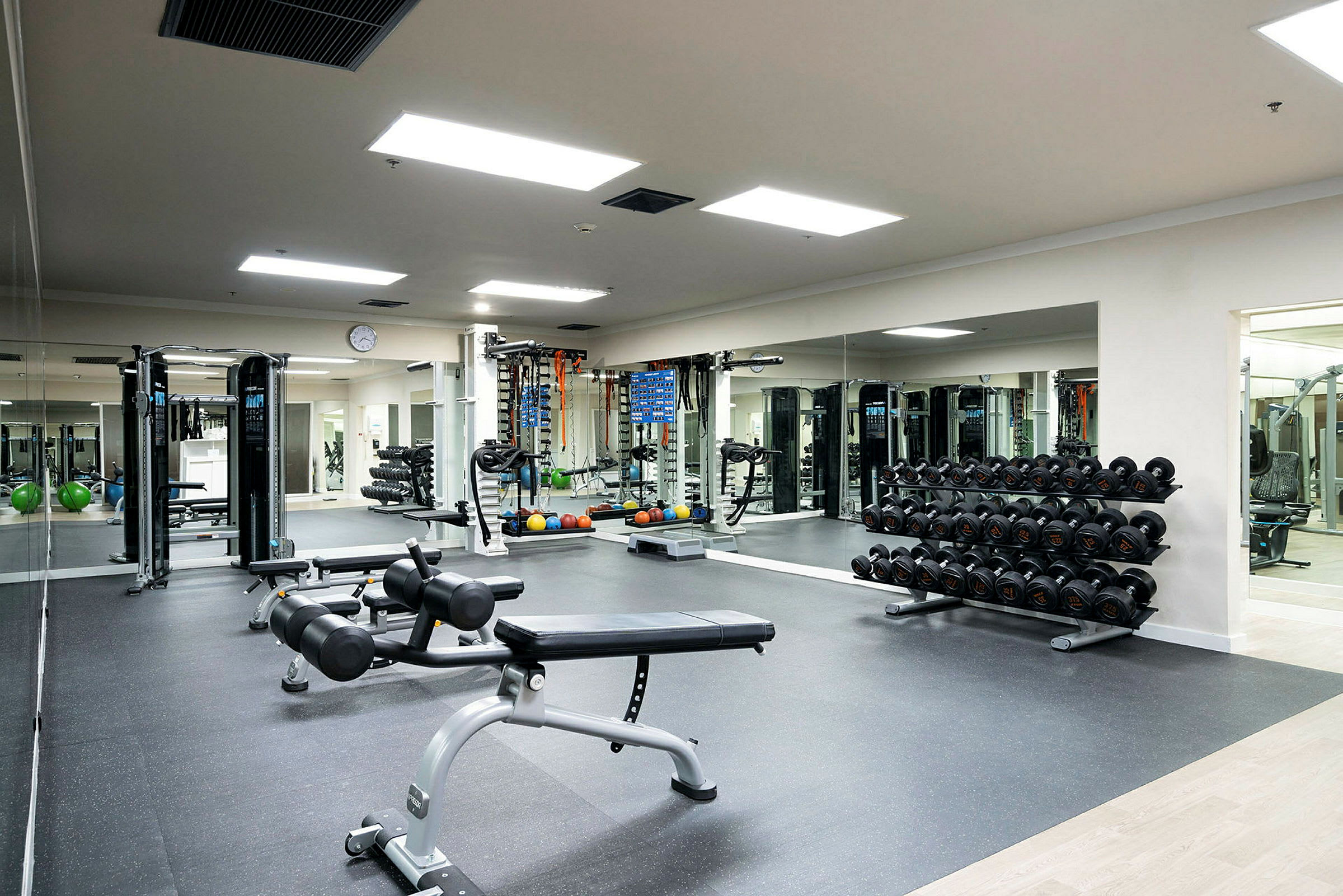 Ett gym med fria vikter, träningsmaskiner och träningsbänkar, med spegelväggar och ljusa tak i bakgrunden. (AI-genererad text)