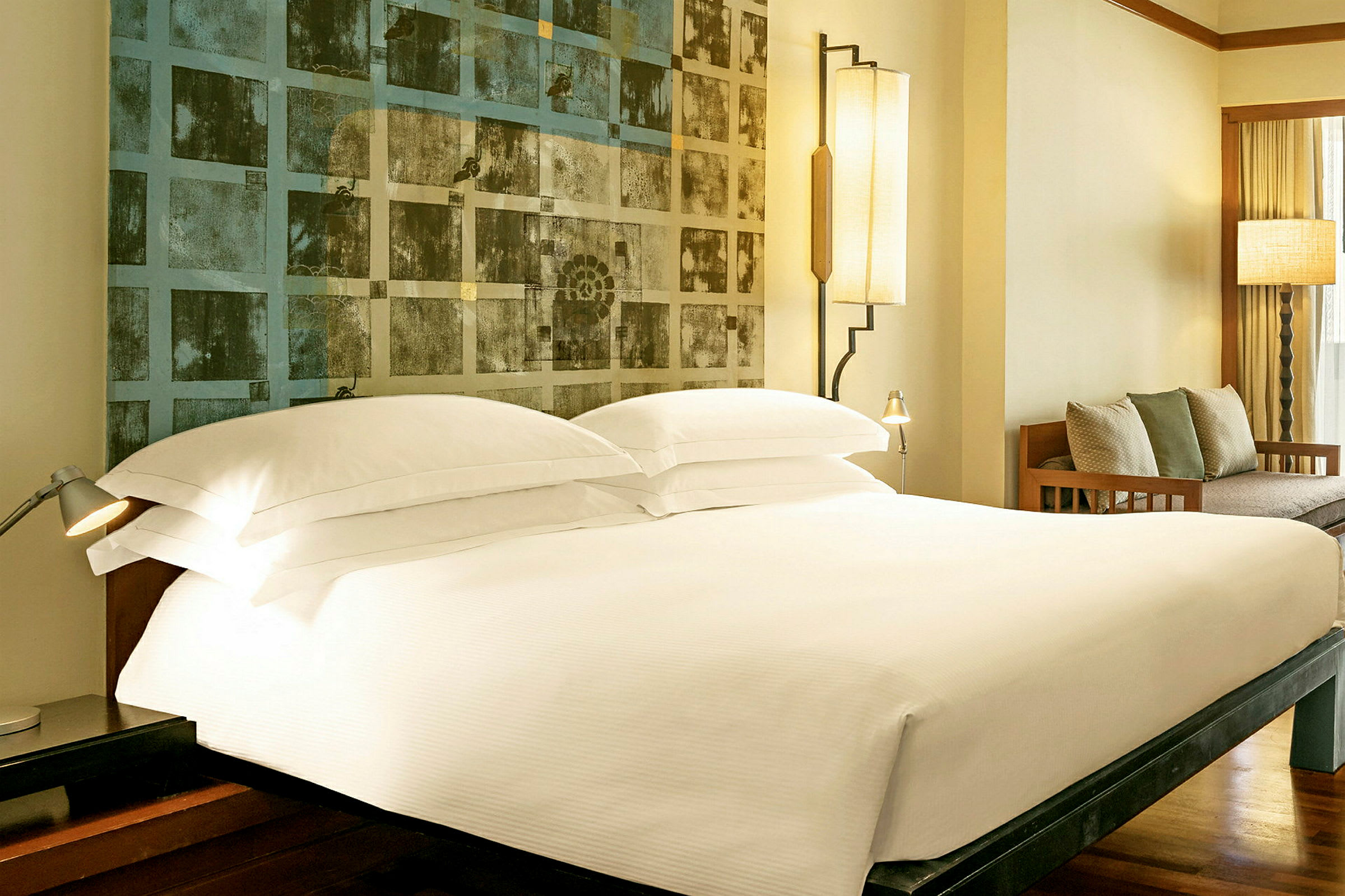 Ett hotellrum med en dubbelsäng, inrett med vita sängkläder, två läslampor och en modern konstprydnad vid huvudgaveln. I bakgrunden finns en soffa med prydnadskuddar. (AI-genererad text)