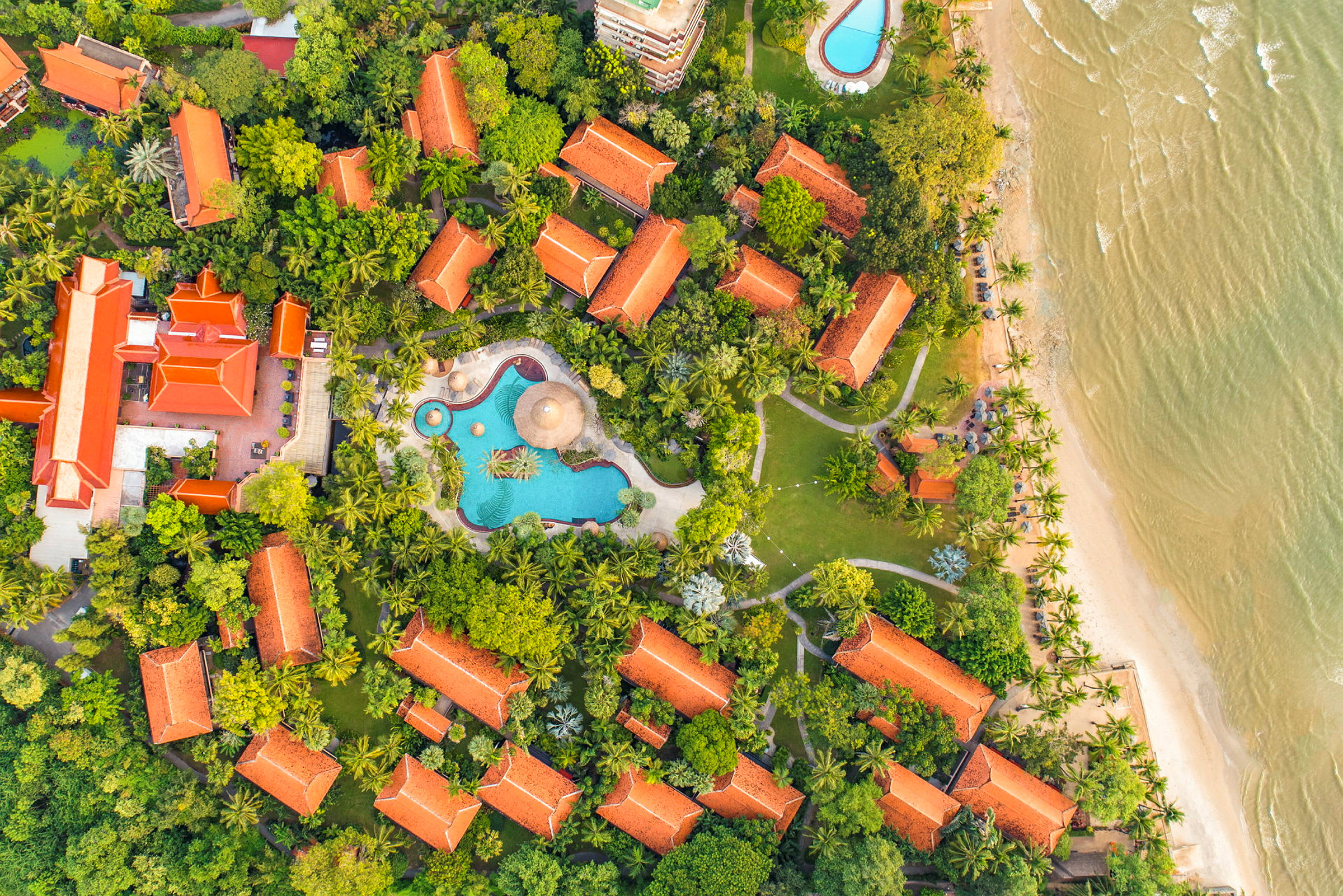 Flygbild över en resort med röda tak omringad av grönska, simbassänger och nära en strand. (AI-genererad text)