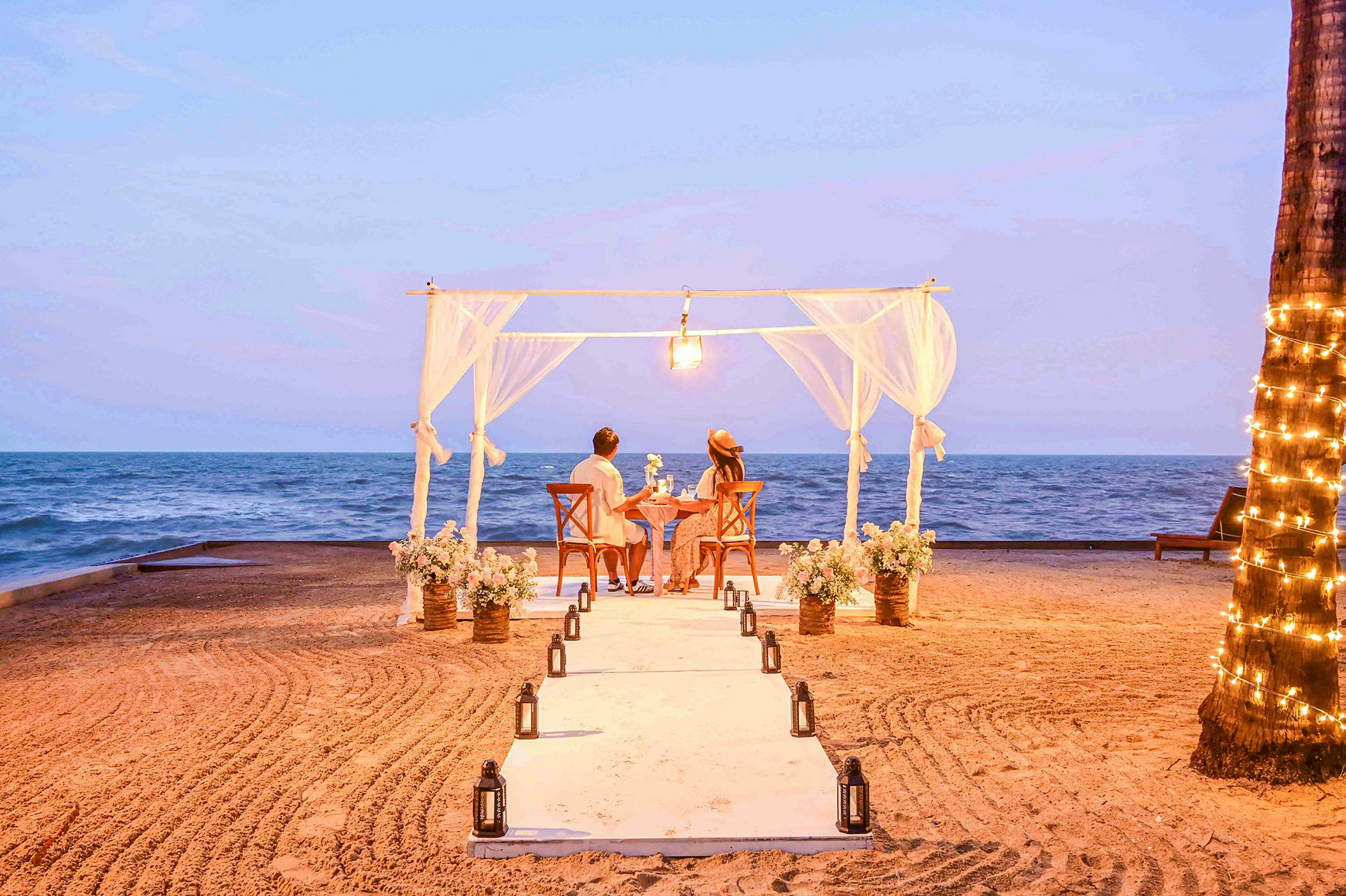 Ett par sitter vid ett dukat bord under ett tält dekorerat med tyger och blommor på en strand framför havet. (AI-genererad text)