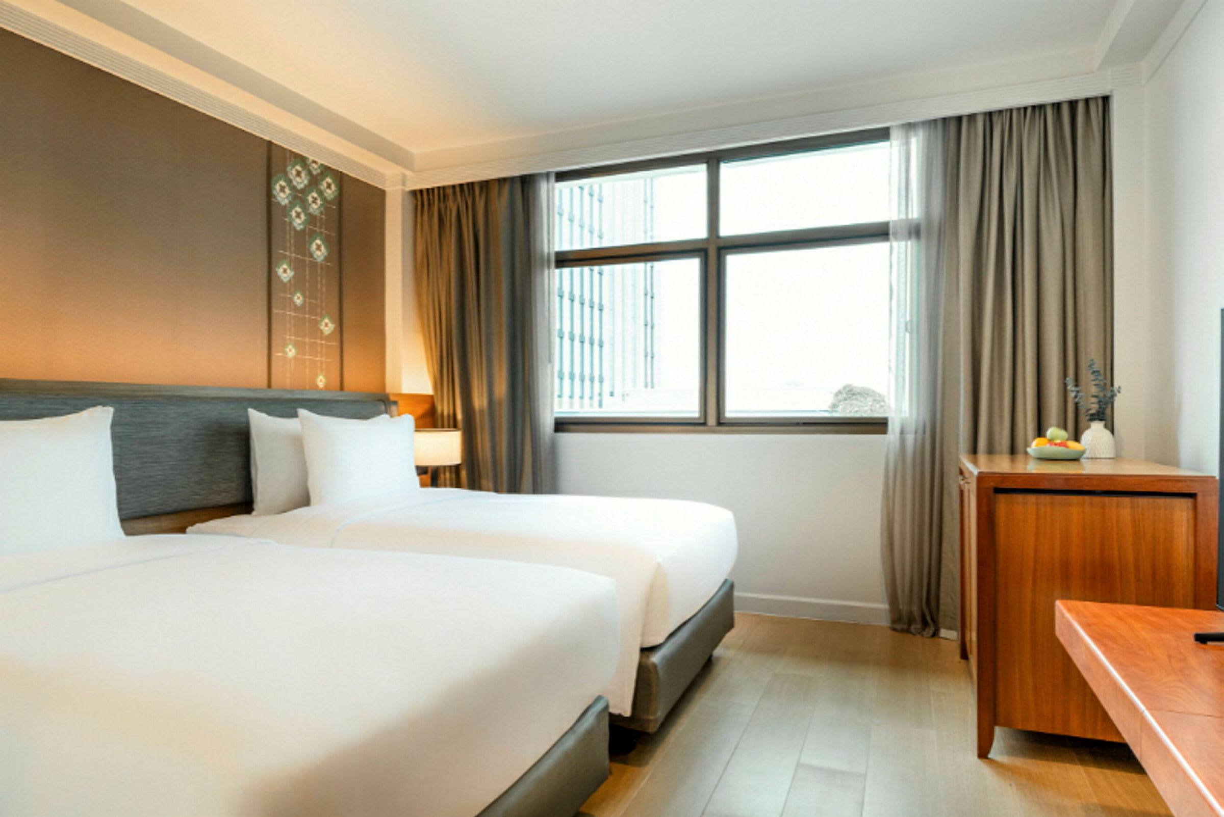 Ett hotellrum med två enkelsängar, en stor fönsterruta med gardiner, samt en träbyrå med en skål med frukt och dekoration. (AI-genererad text)