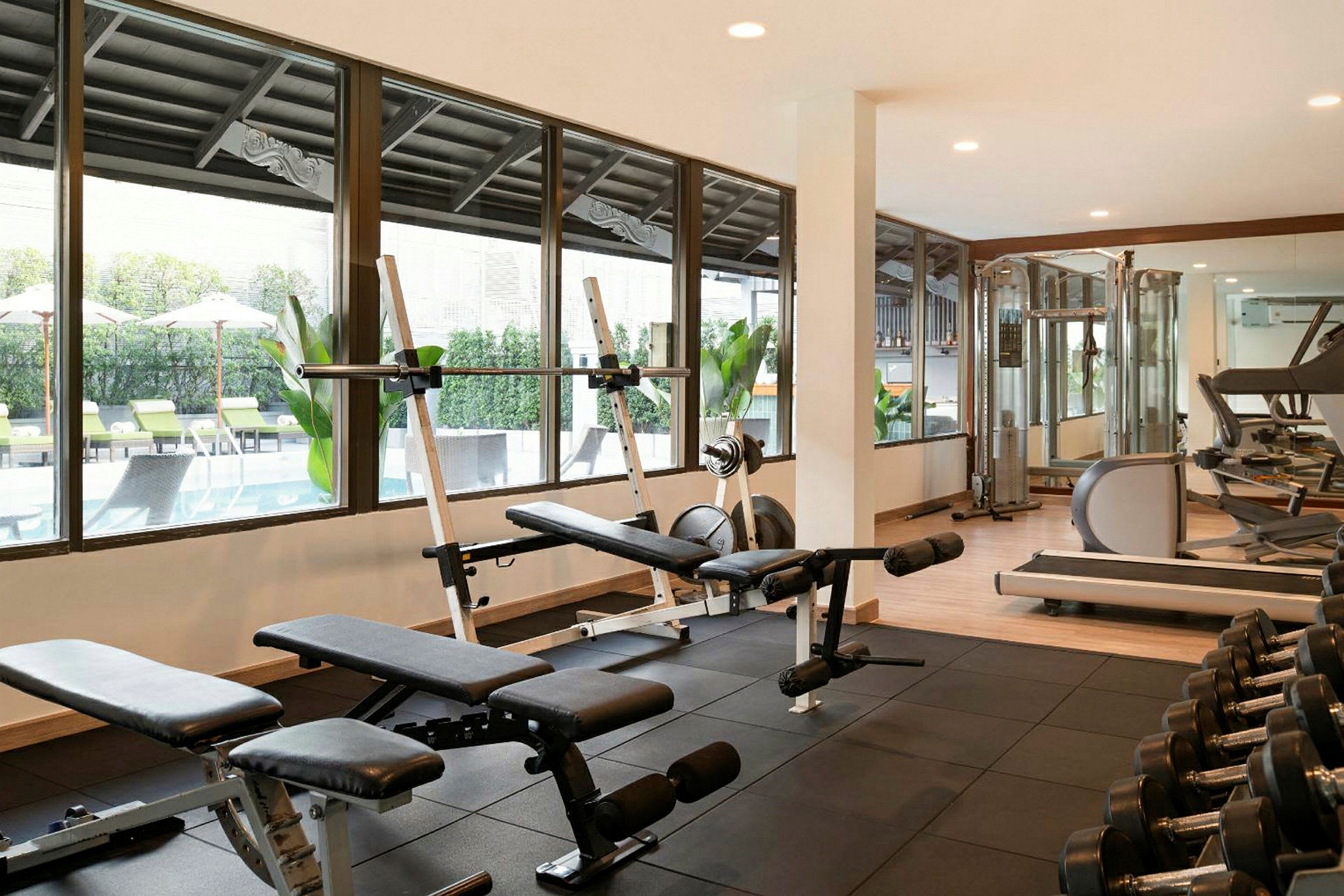 Ett gym med träningsmaskiner och viktbänkar, fönster som vetter mot en pool utomhus. (AI-genererad text)
