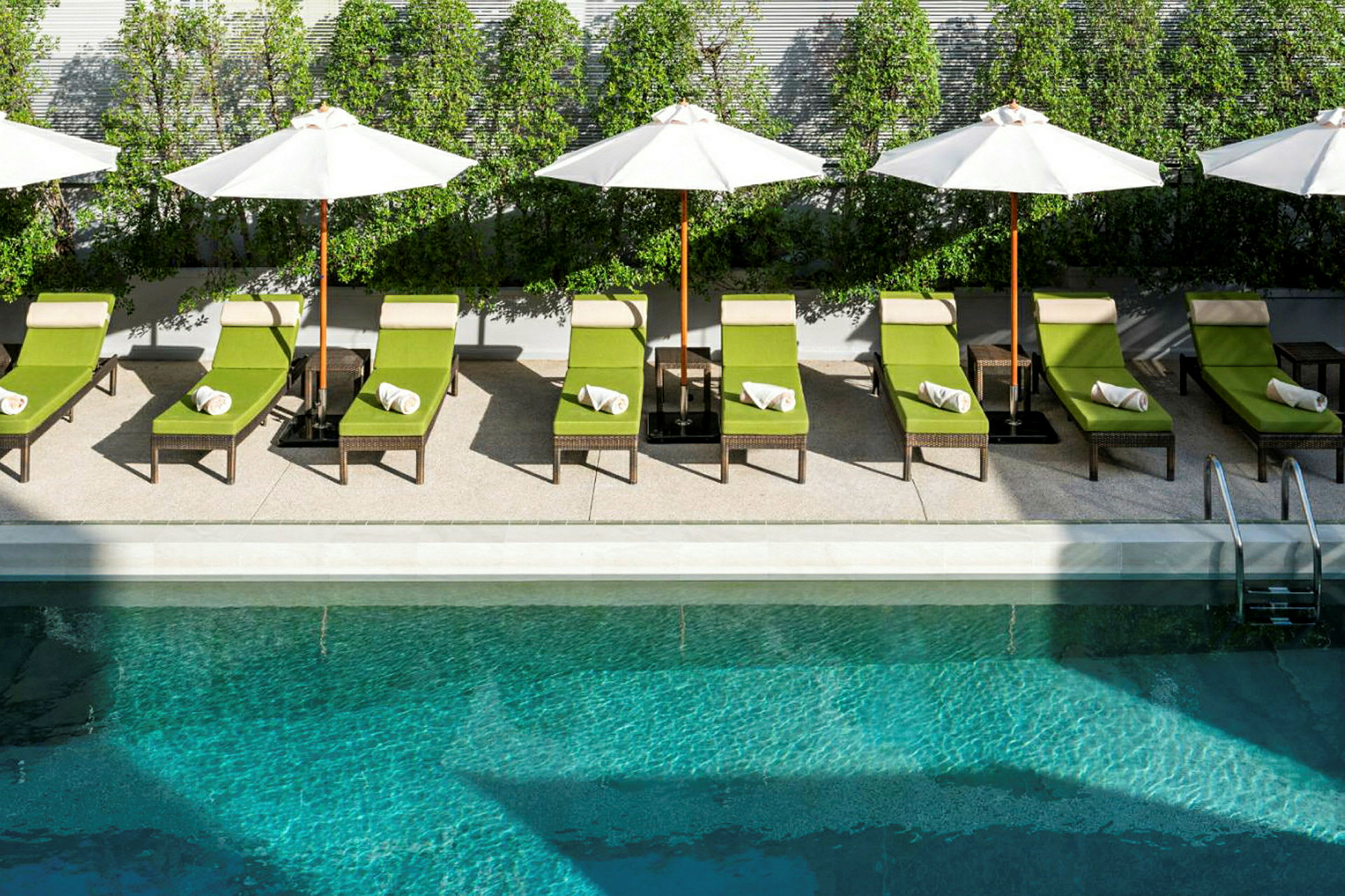 Flera solstolar med gröna dynor och vita handdukar står uppradade vid sidan av en pool. Paraplyer ger skugga, och gröna buskar är i bakgrunden. (AI-genererad text)