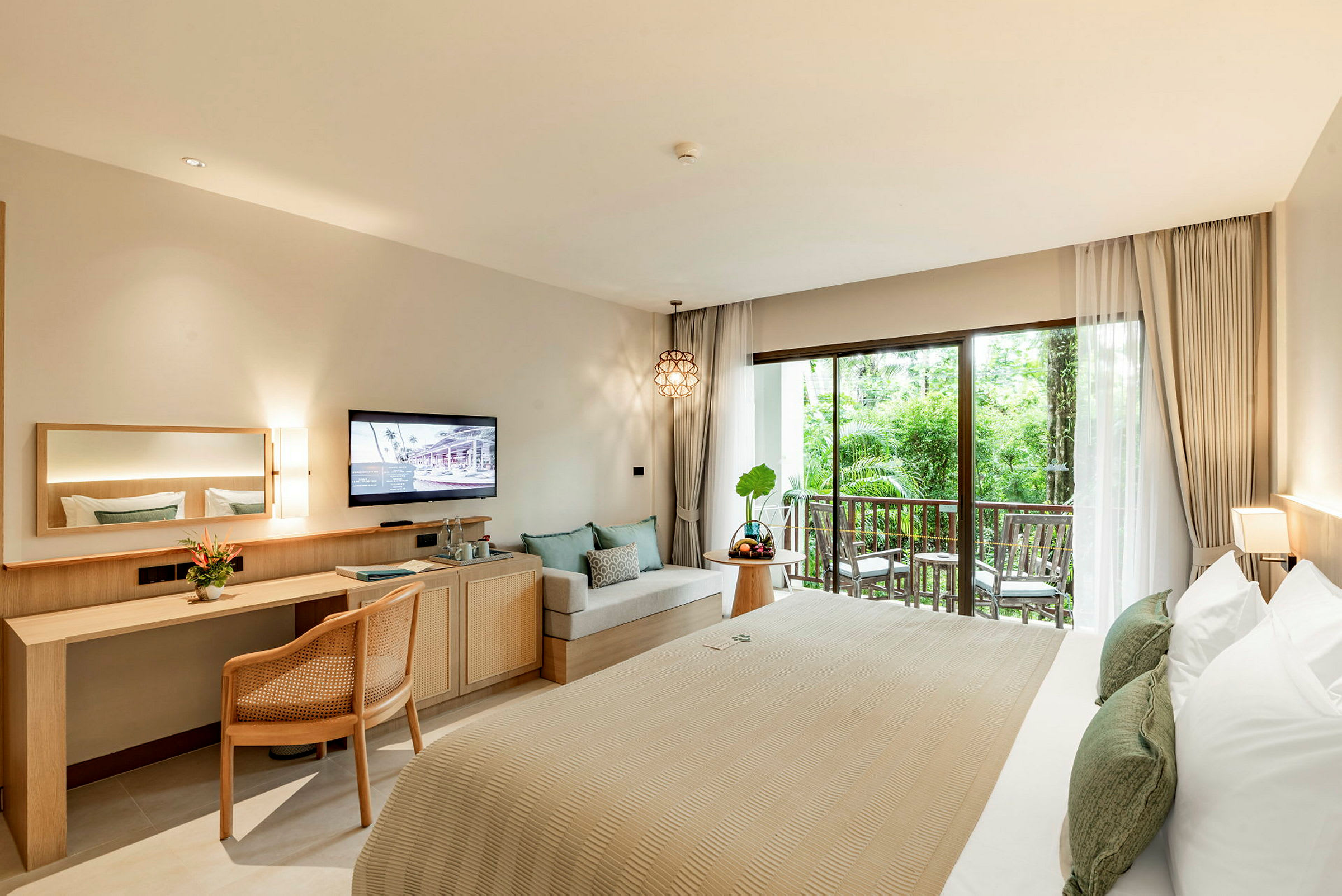 Ett hotellrum med en säng, ett skrivbord, en TV, en sittgrupp och en balkong med utsikt över grönska. (AI-genererad text)