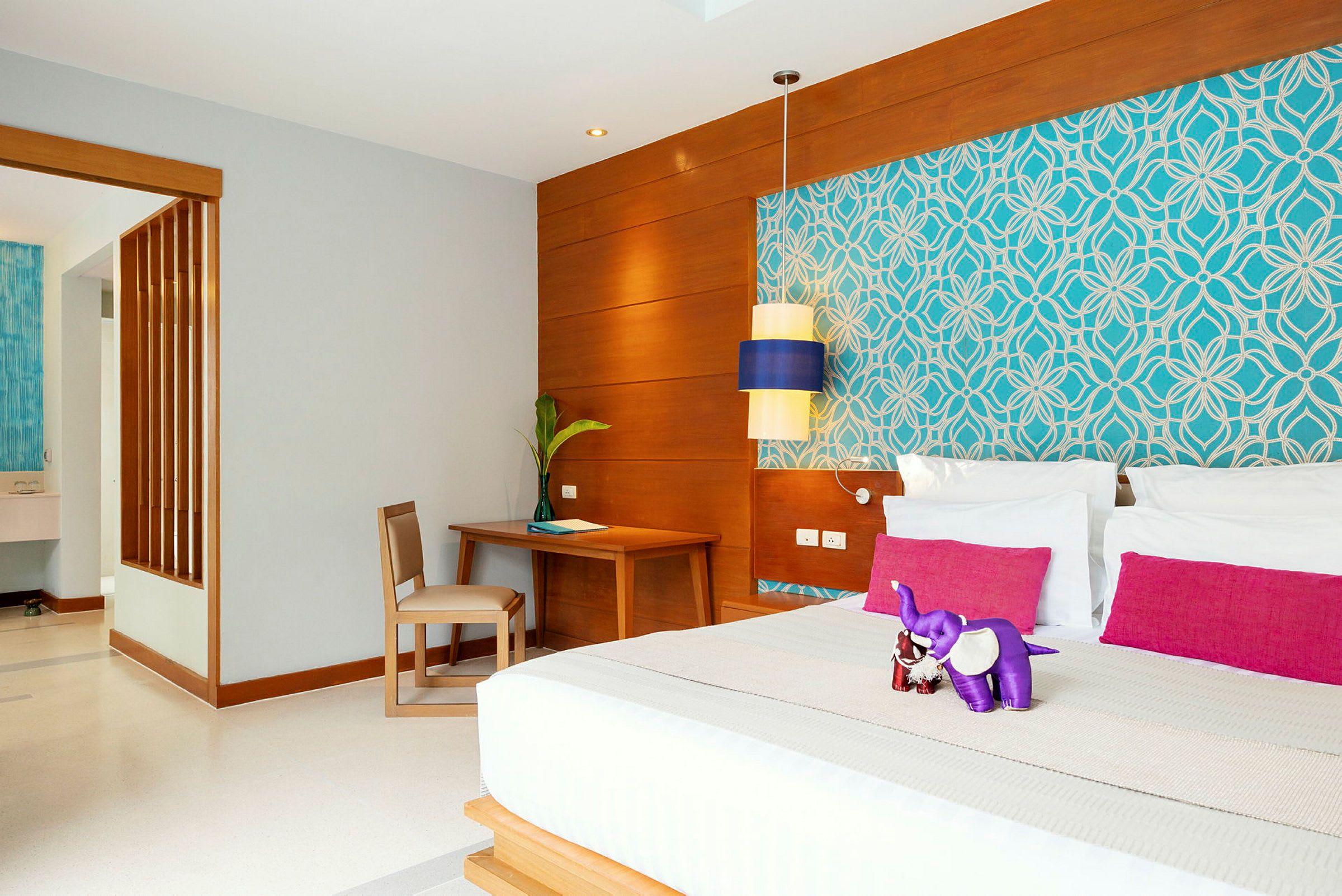Ett hotellrum med en stor säng, rosa kuddar, och en lilafärgad elefantprydnad. Modern inredning med trädetaljer och blommönstrad tapet. (AI-genererad text)