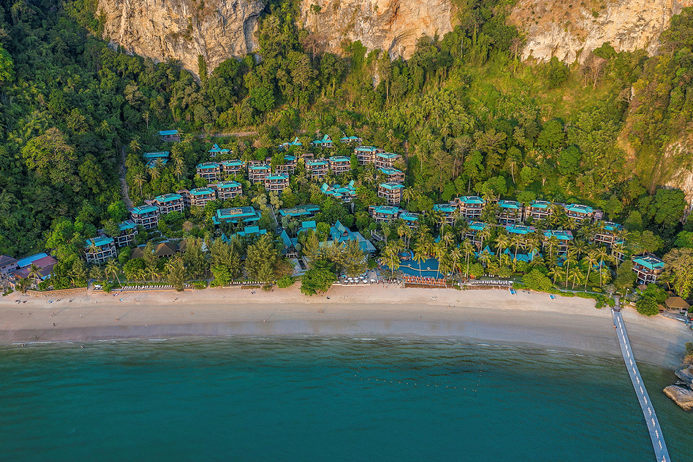 Flygfoto av en strand med turkost vatten, hotellbyggnader i naturmiljö och omgivande grönska. (AI-genererad text)