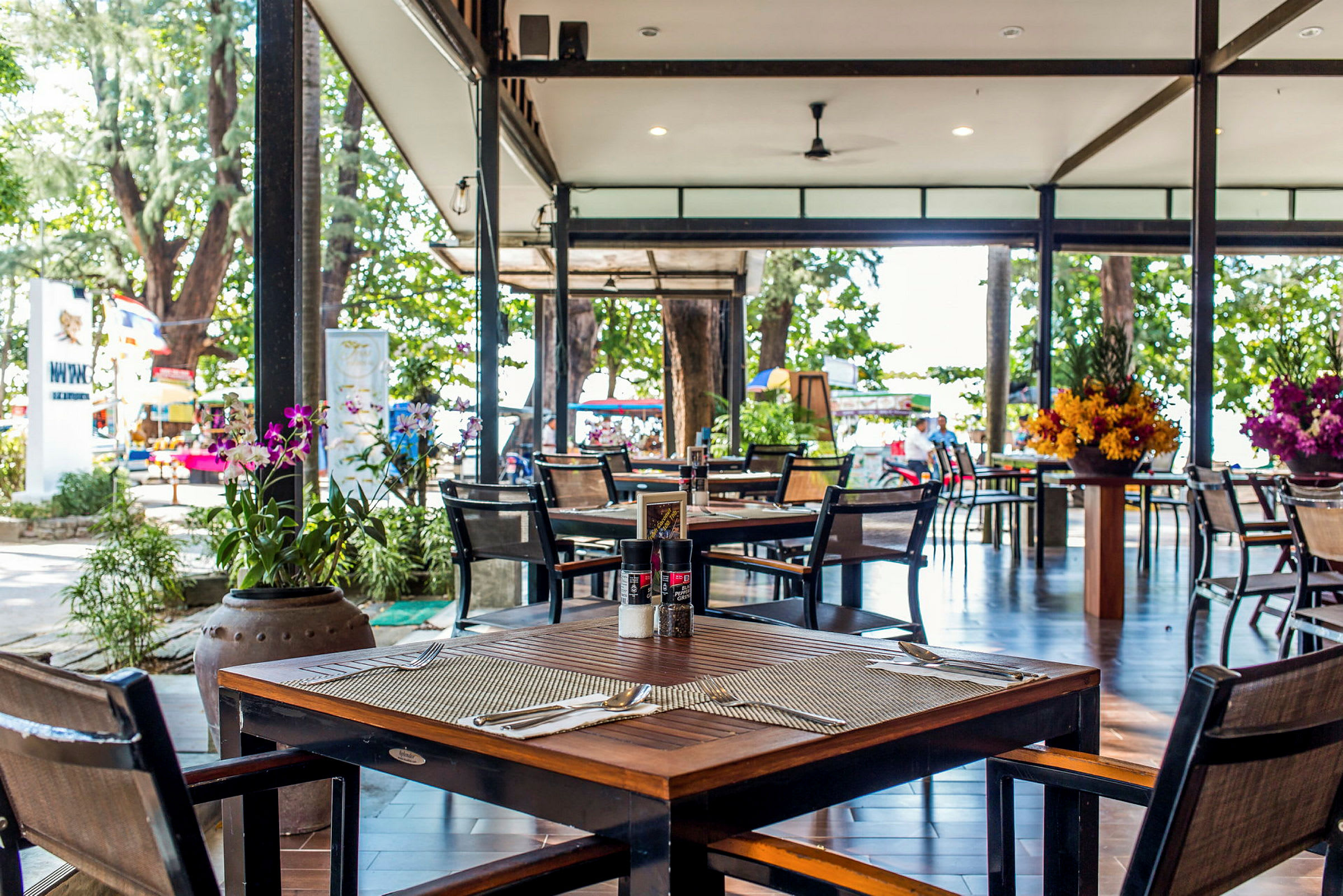 En utomhusrestaurang med möblerade bord och stolar. Stora blomarrangemang pryder omgivningen, och i bakgrunden syns gröna träd och en strand. (AI-genererad text)