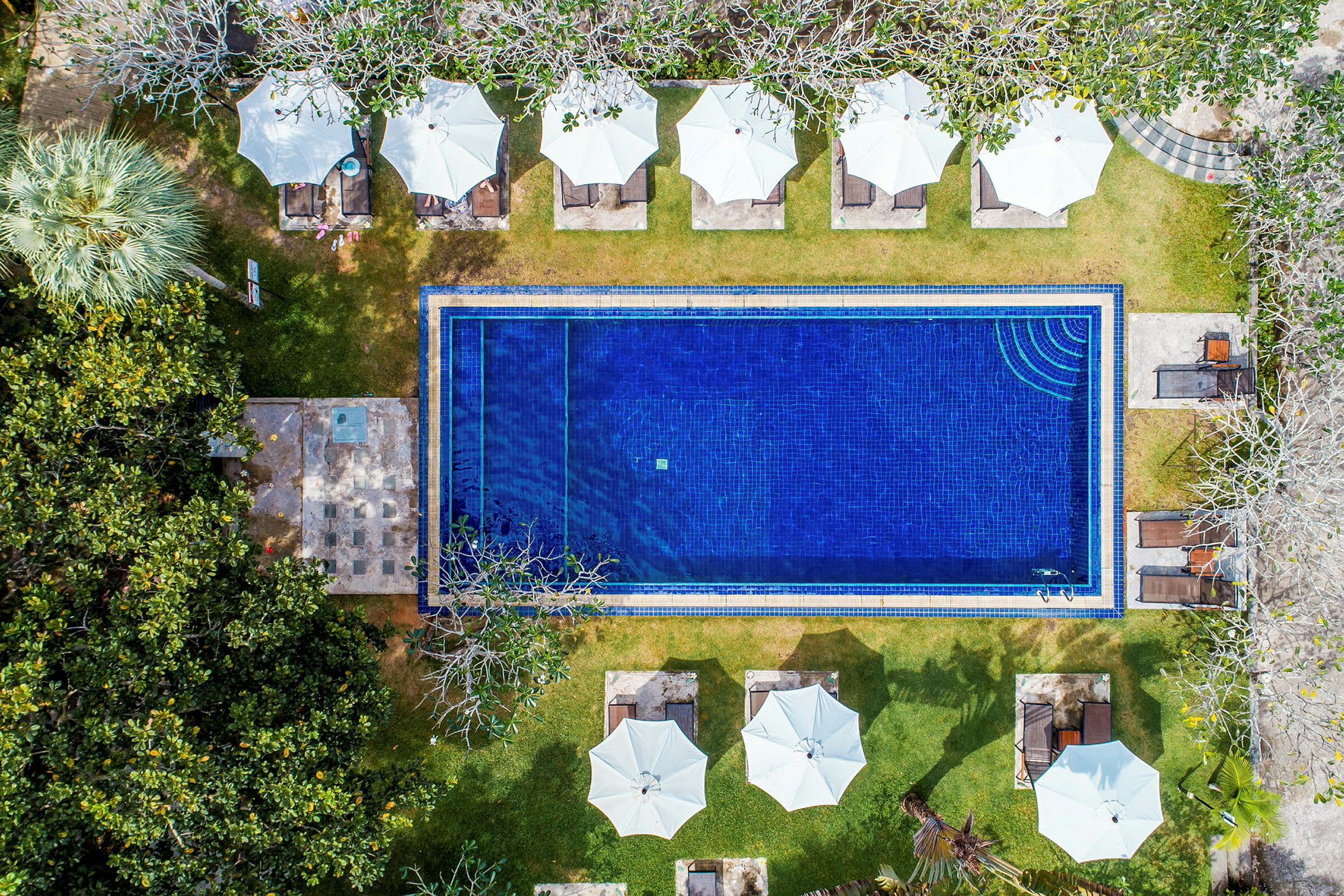 Flygfoto över en rektangulär pool omgiven av grönska, solstolar och vita parasoller. (AI-genererad text)
