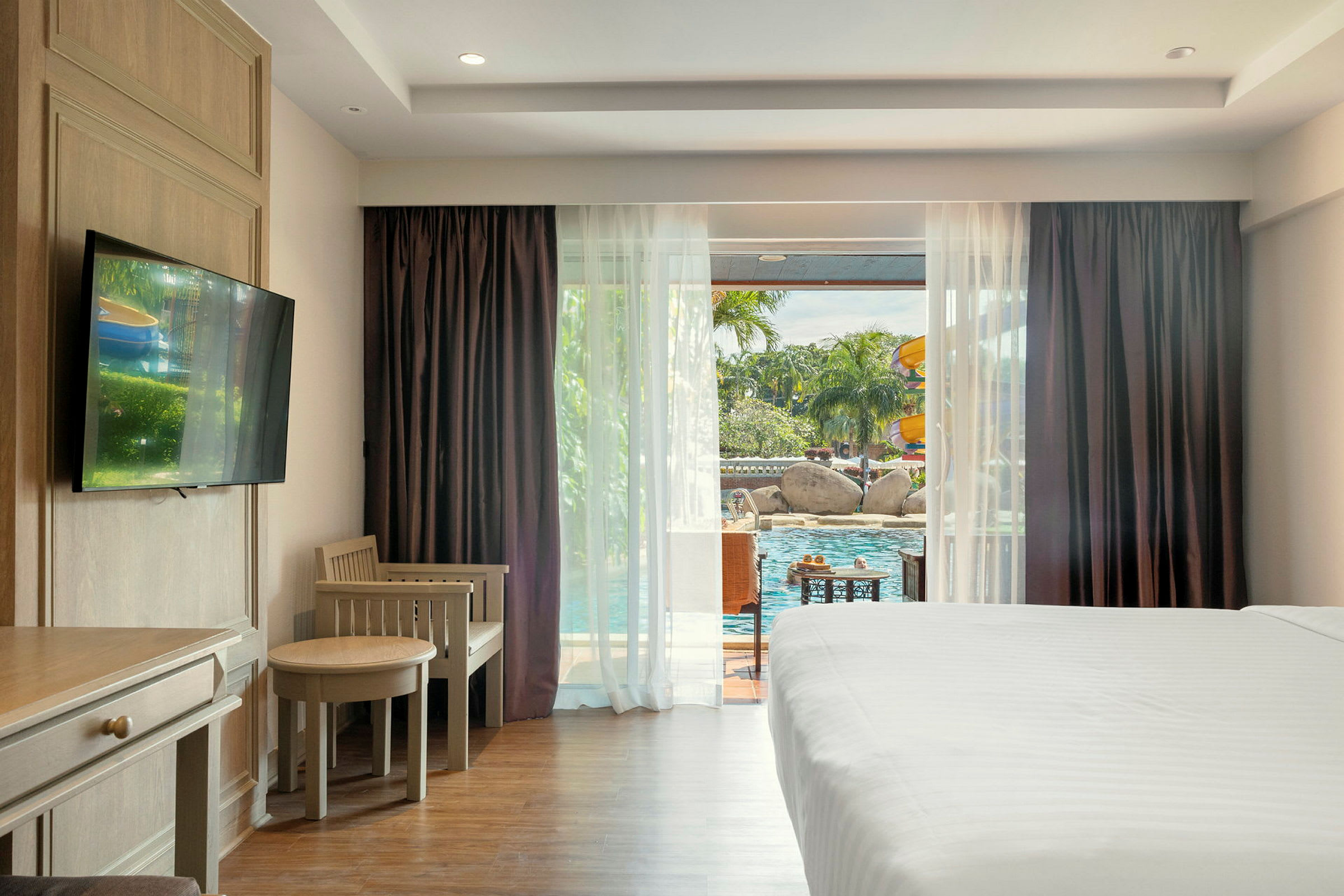 Ett hotellrum med ljusa möbler, en säng och utsikt över en pool utanför genom stora fönster. (AI-genererad text)