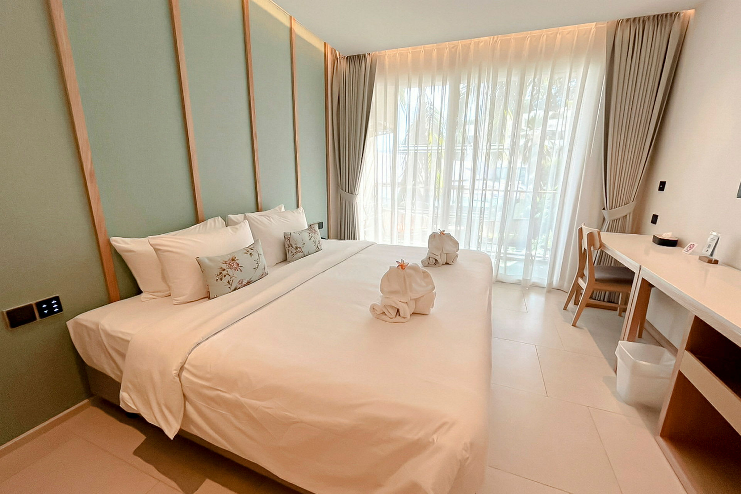 Bild av ett modernt hotellrum med en stor bäddad säng, vita sängkläder och dekorativa kuddar, samt handdukar formade som svanar. Rummet har fönster med ljusa gardiner och en skrivbordsplats vid sidan av. (AI-genererad text)