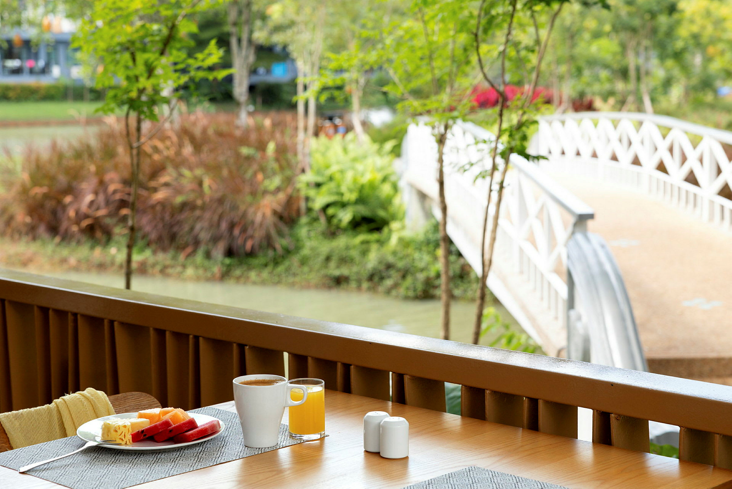 Frukostbord med kaffe och juice på en terrass med utsikt mot en vit bro som leder över en kanal. (AI-genererad text)