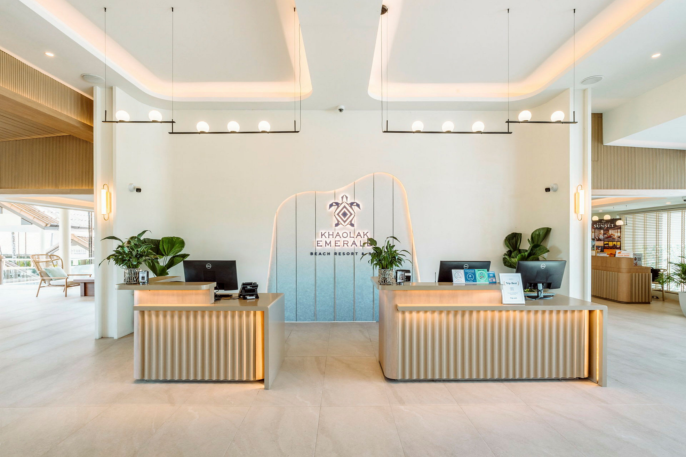 Receptionområde med två moderna skrivbord dekorerade med växter. En logotyp med texten 'Khao Lak Emerald Beach Resort' är placerad på väggen. (AI-genererad text)