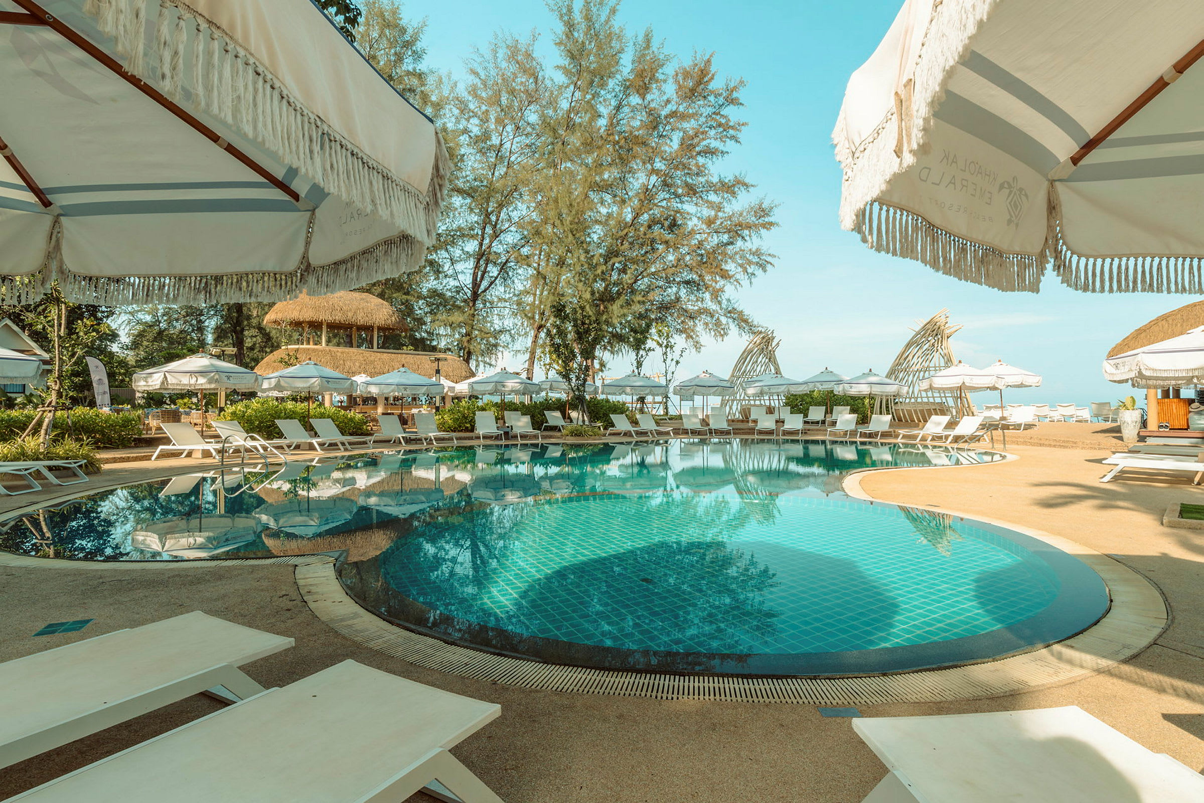 Poolområde vid en resort med parasoller och solstolar, omgiven av träd och utsikt mot havet. (AI-genererad text)