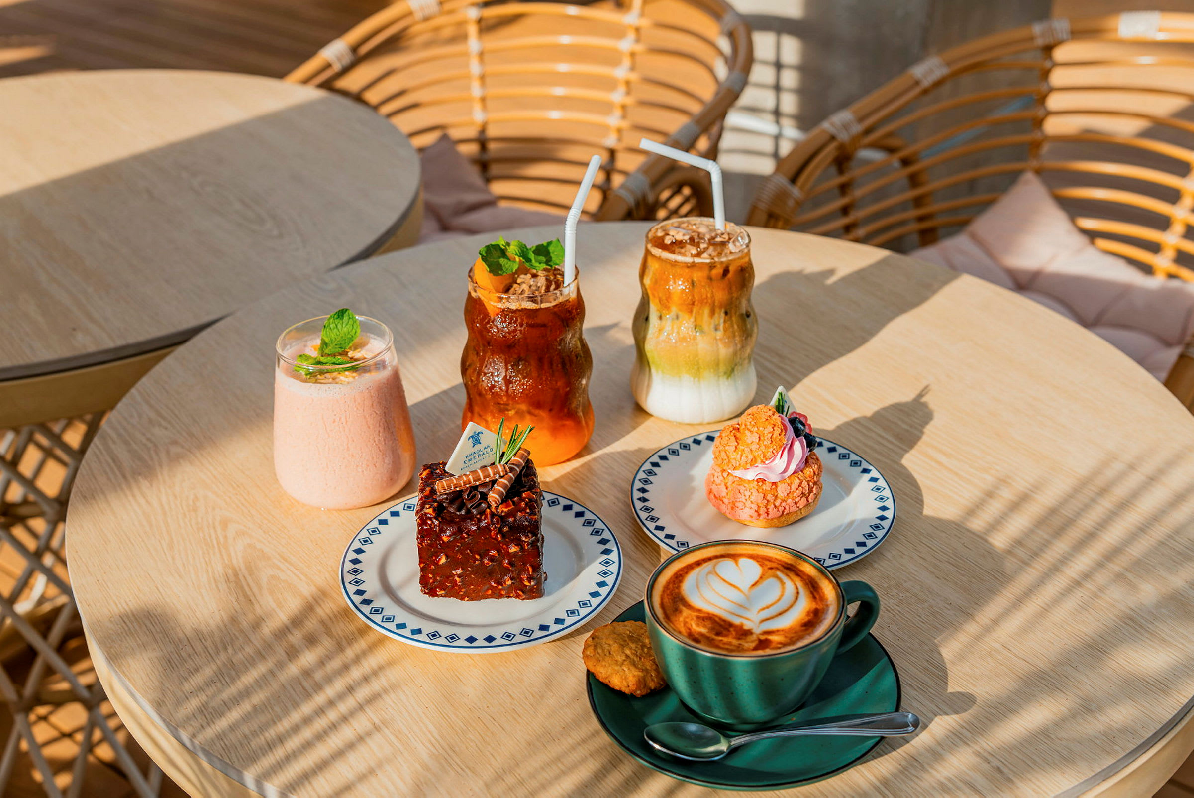 Bilden visar ett bord med olika bakverk och drycker. Det finns en kopp kaffe med latte art, en brownie, ett rosa bakverk samt några kalla drycker. (AI-genererad text)