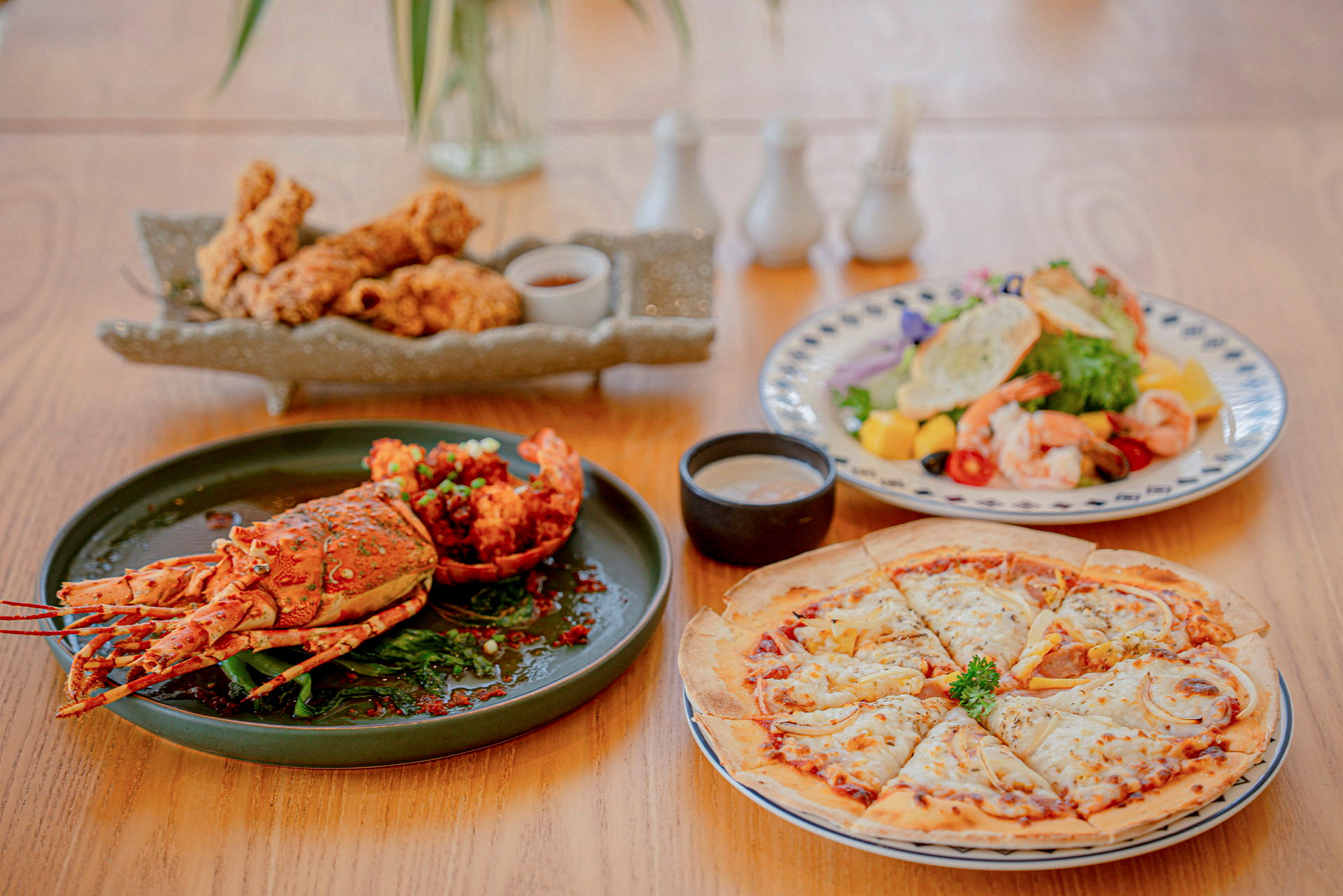 Ett matbord med rätter som inkluderar en hummer, en tallrik med friterad kyckling, en sallad med räkor och tropiska frukter, samt en pizza. (AI-genererad text)