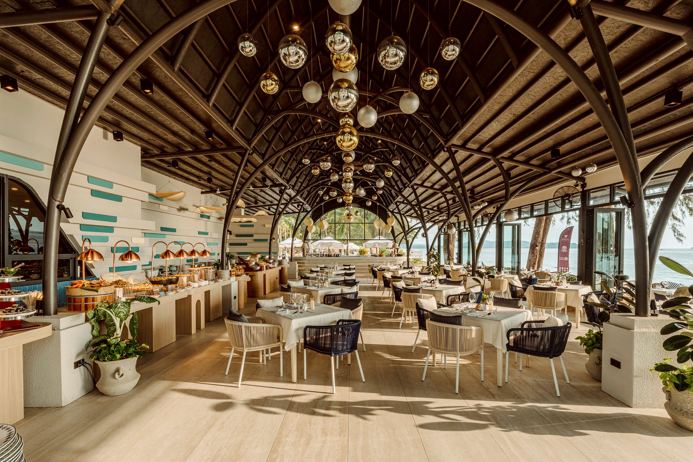 En restaurang med vacker arkitektur och takdekorationer, fylld med dukade bord och en buffédisk. Inkluderar utsikt över havet i bakgrunden. (AI-genererad text)