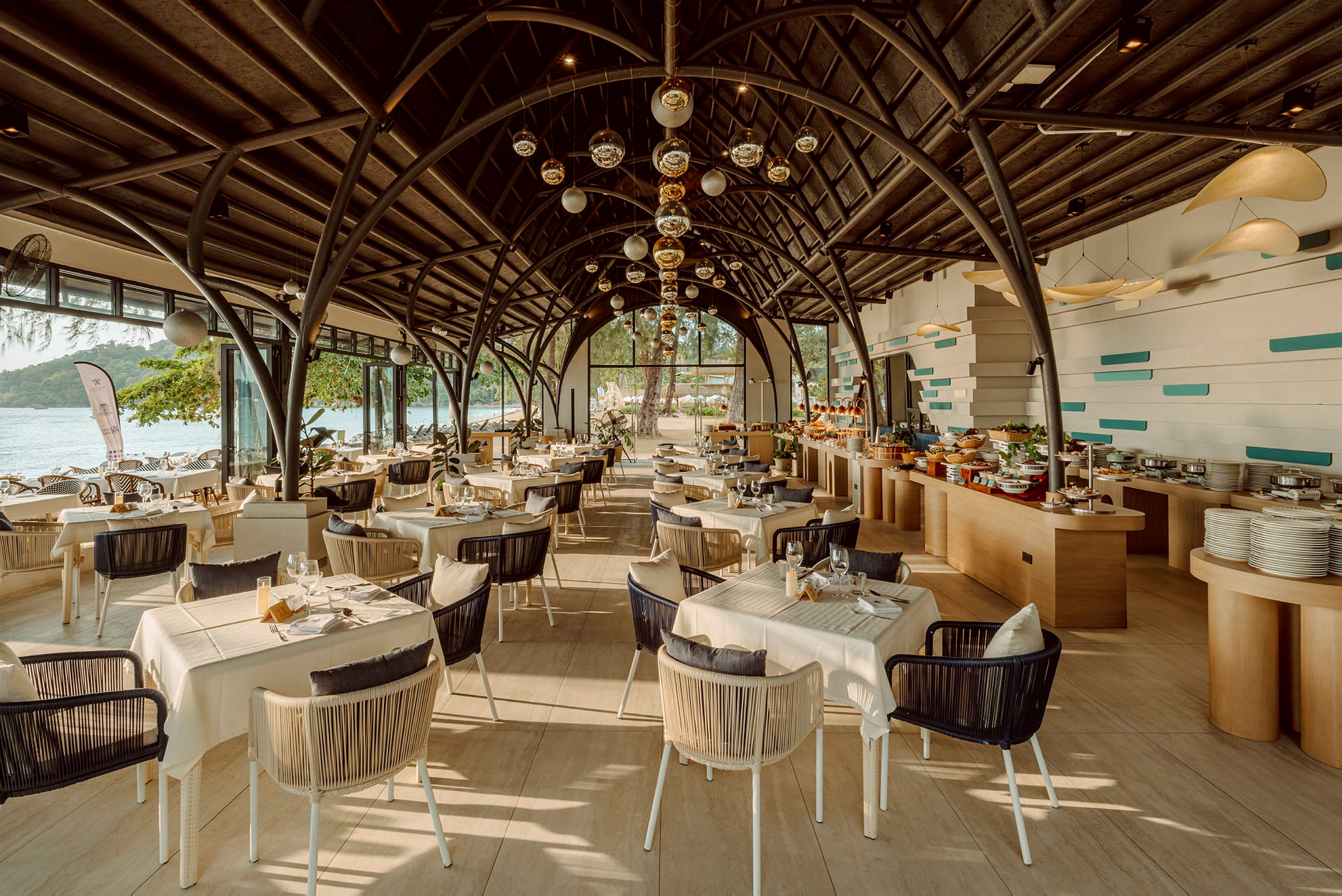 En modern restaurang med stora fönster som vetter mot havet. Bord och stolar är snyggt arrangerade under en välvd takstruktur, och det finns en buffé till höger. (AI-genererad text)