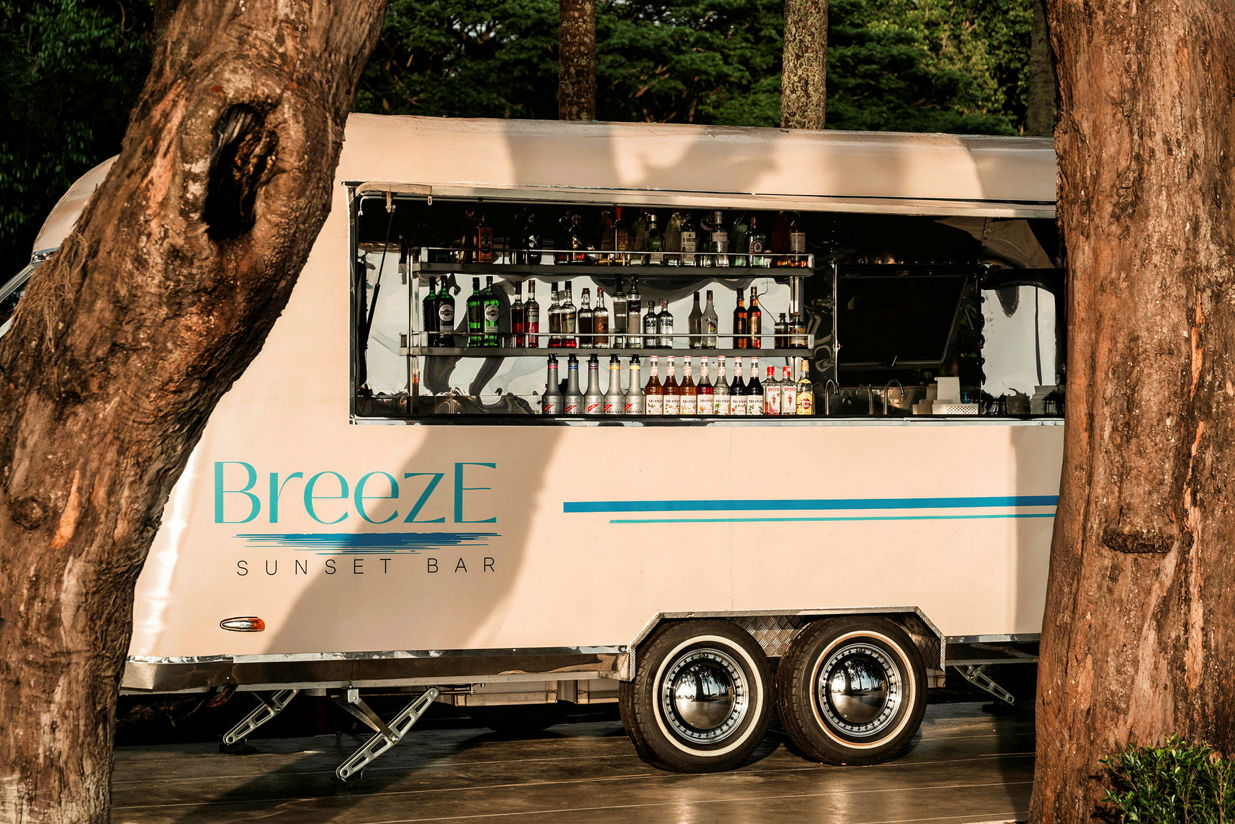 En mobil bardel med märket 'BreezE Sunset Bar' är uppställd mellan träden, med flaskor och drycker synliga i det öppna serviceområdet. (AI-genererad text)