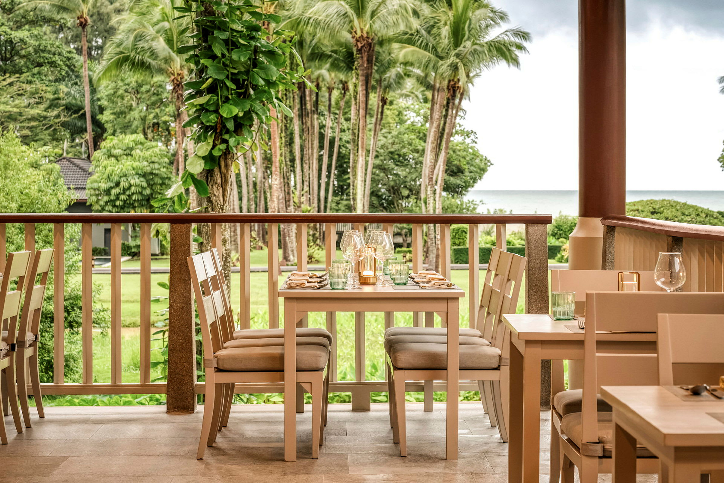 En restaurang med bord och stolar står på en veranda med utsikt över en grön trädgård och palmer, samt havet i bakgrunden. (AI-genererad text)