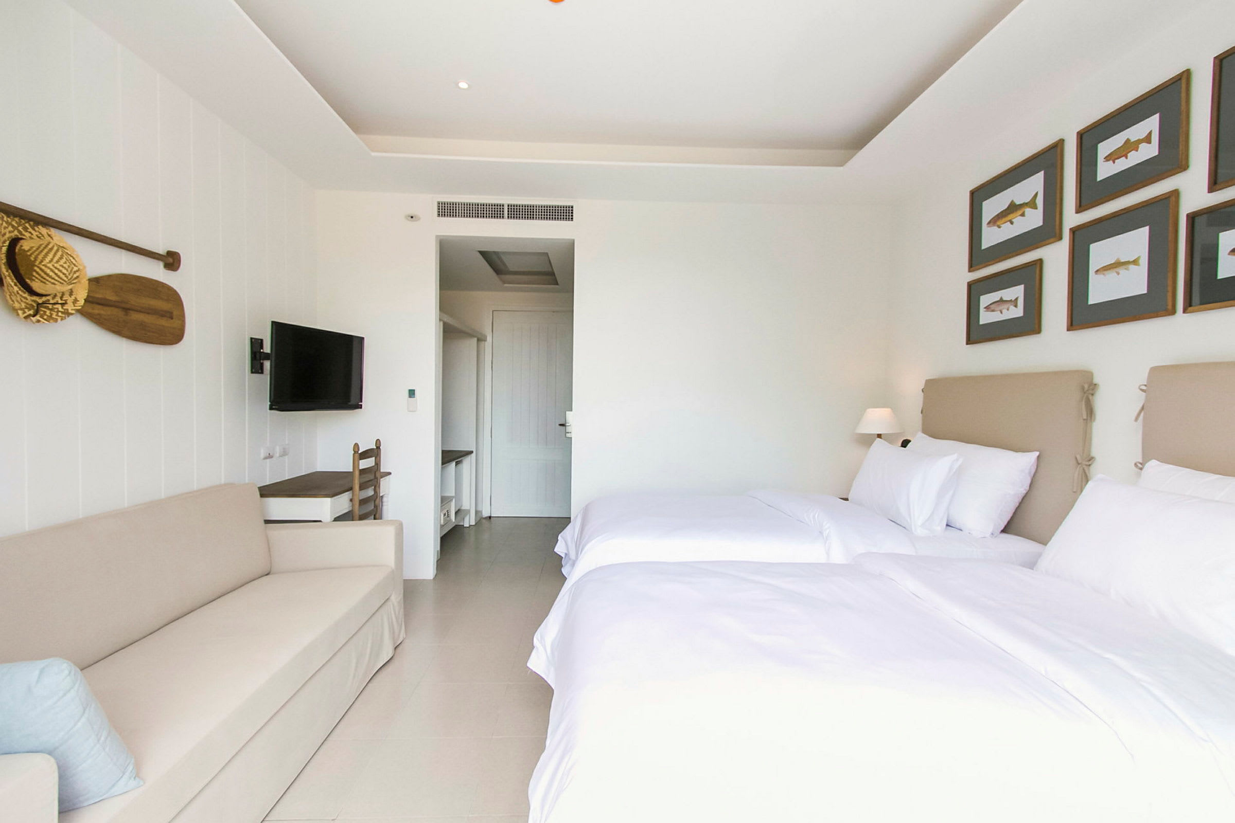 Ett ljust hotellrum med två sängar bäddade med vita lakan, en beige soffa och en väggmonterad TV. Väggdekorationen inkluderar fiskmotiv i ramar och en träpaddel med en stråhatt. (AI-genererad text)