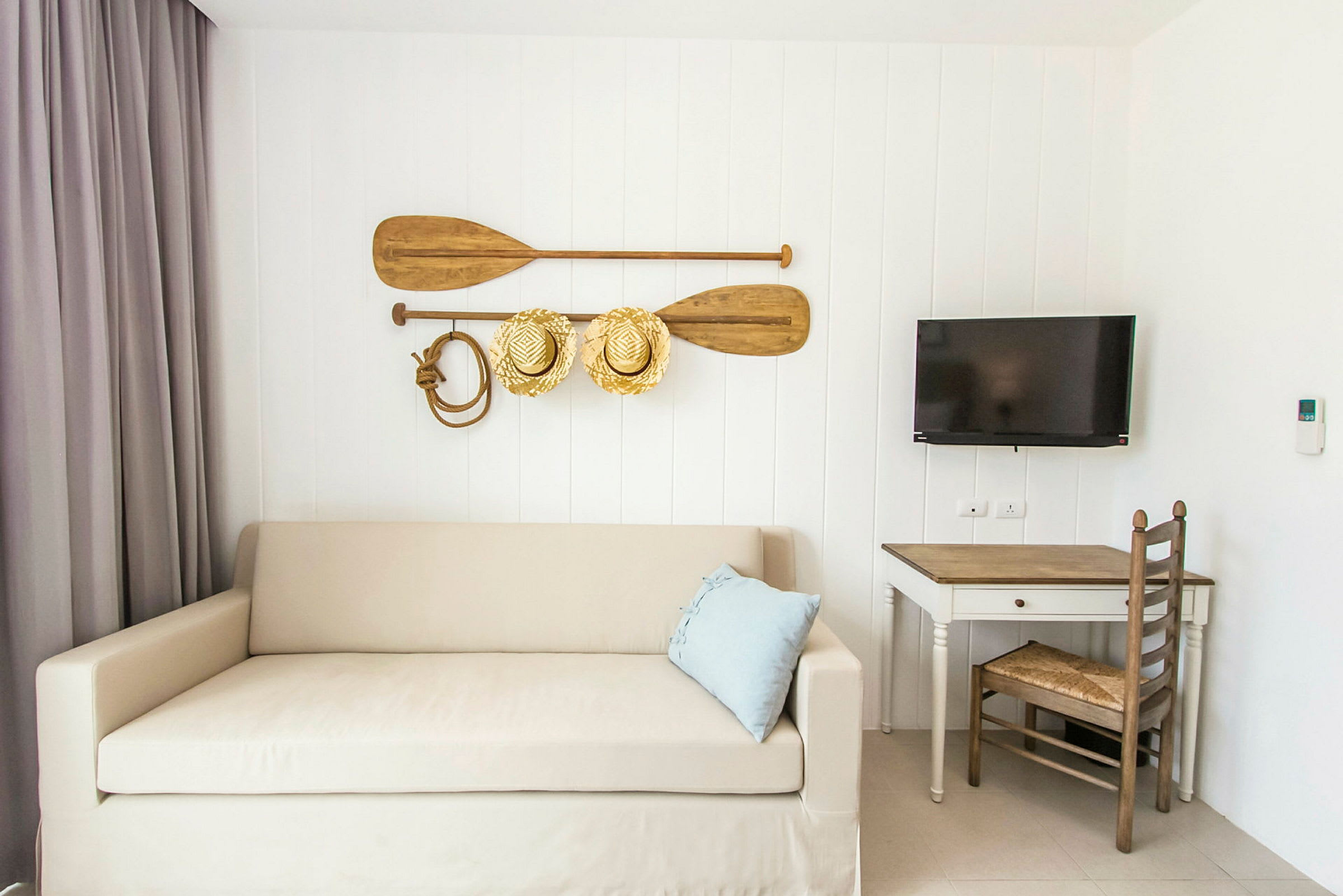 Ett rum med en beige soffa, blå kudde och väggdekorationer med åror och hattar. Vid sidan finns ett skrivbord med stol och en väggmonterad TV. (AI-genererad text)