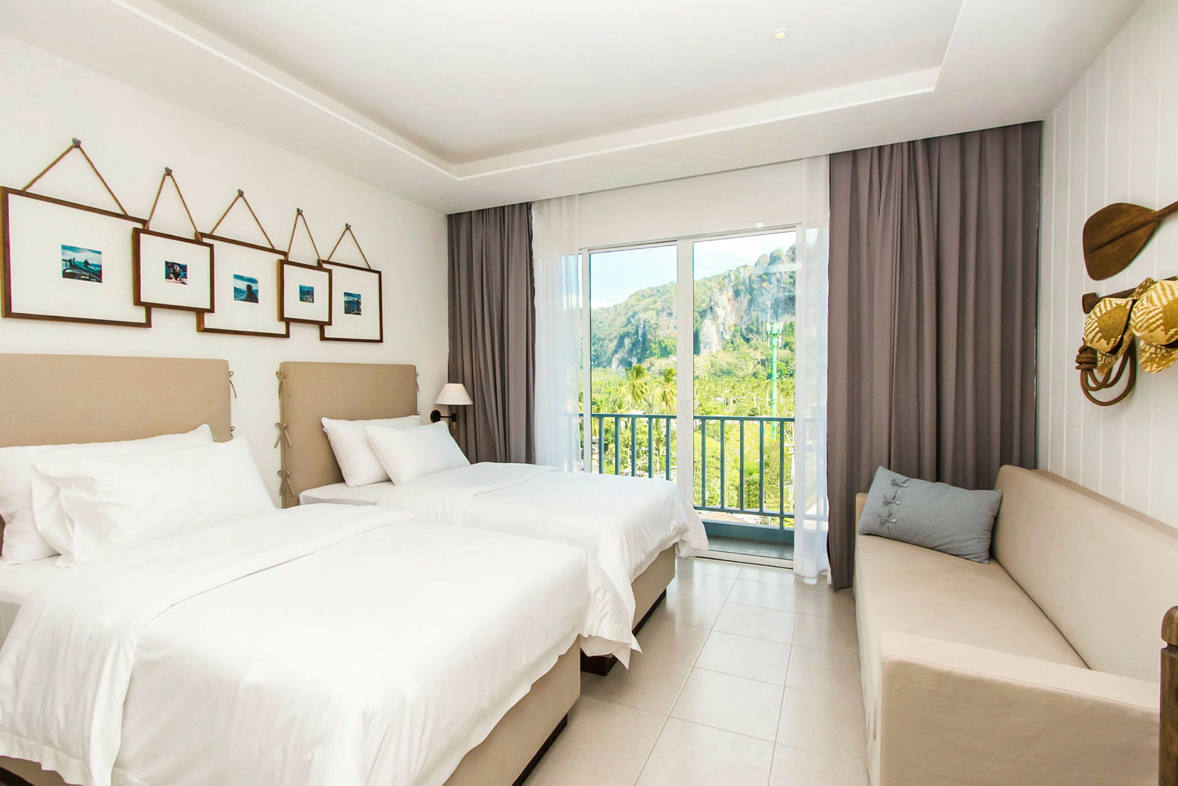 Ett hotellrum med två enkelsängar, en soffa och en balkong med utsikt över grönska och klippor. (AI-genererad text)