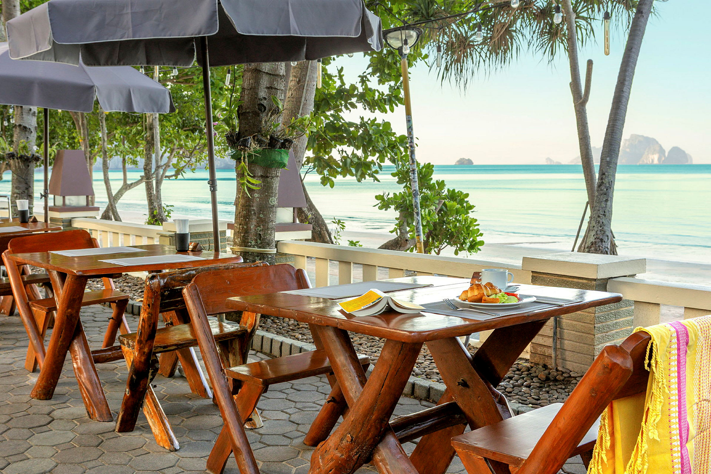 Utsikt från en restaurang vid stranden med träbord, parasoller och havet i bakgrunden. (AI-genererad text)
