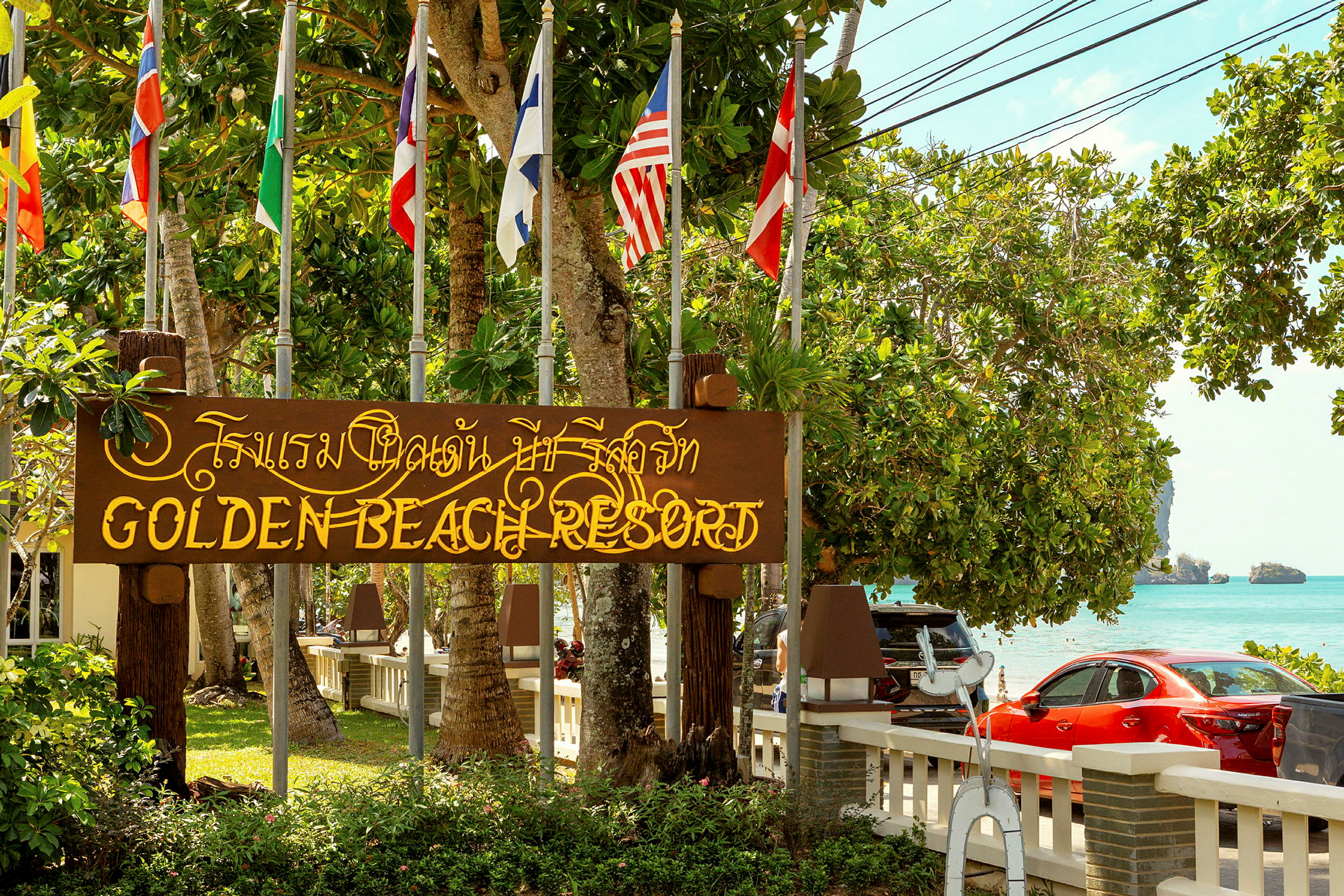En skylt med texten 'Golden Beach Resort' ligger i grönskande omgivning nära havet. Flera flaggor syns ovanför skylten. (AI-genererad text)